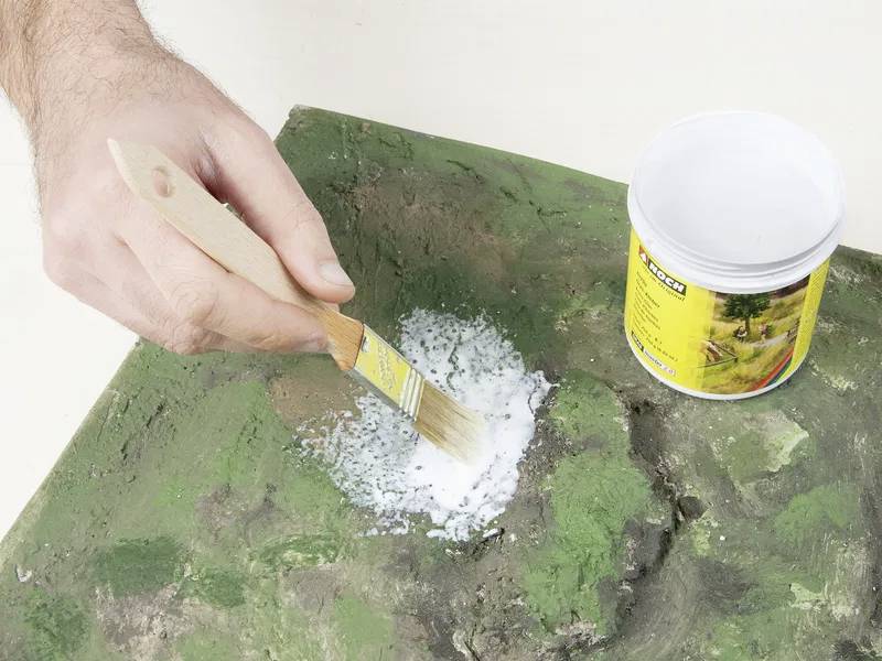 A hand uses a brush to apply a substance from a container onto a green textured surface, possibly for repairing or finishing.