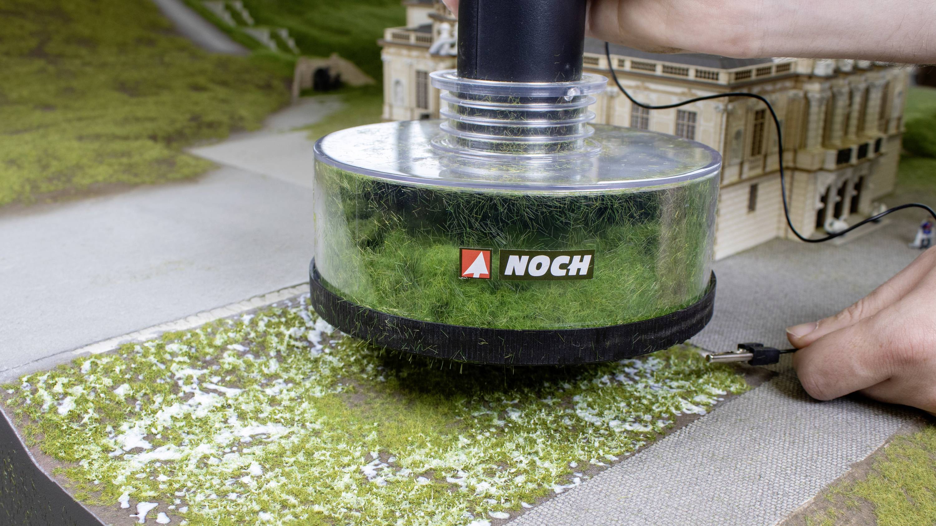 A hand holds a device with 'NOCH' written on it, applying grass to a model of a building complex.