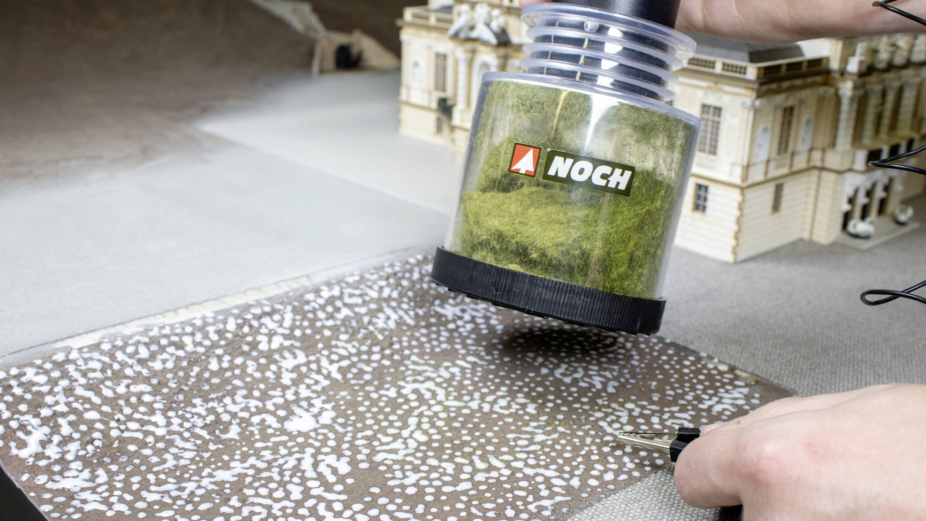A model maker uses a device to distribute grass fibres across a model landscape. In the background, a miniature building stands.