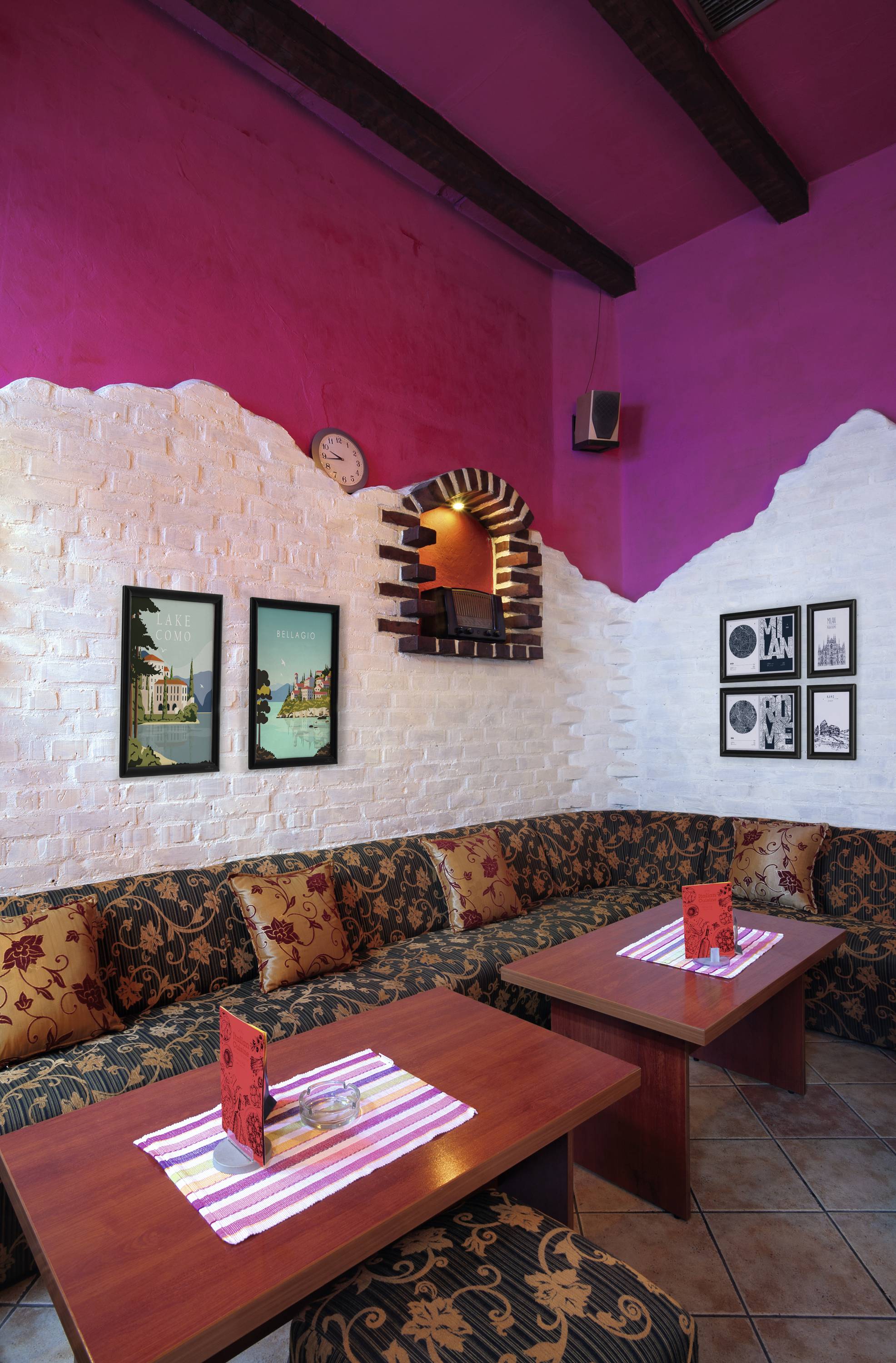 A cosy café with patterned sofas, wooden tables, and decorative posters on a pink and white wall in the background.