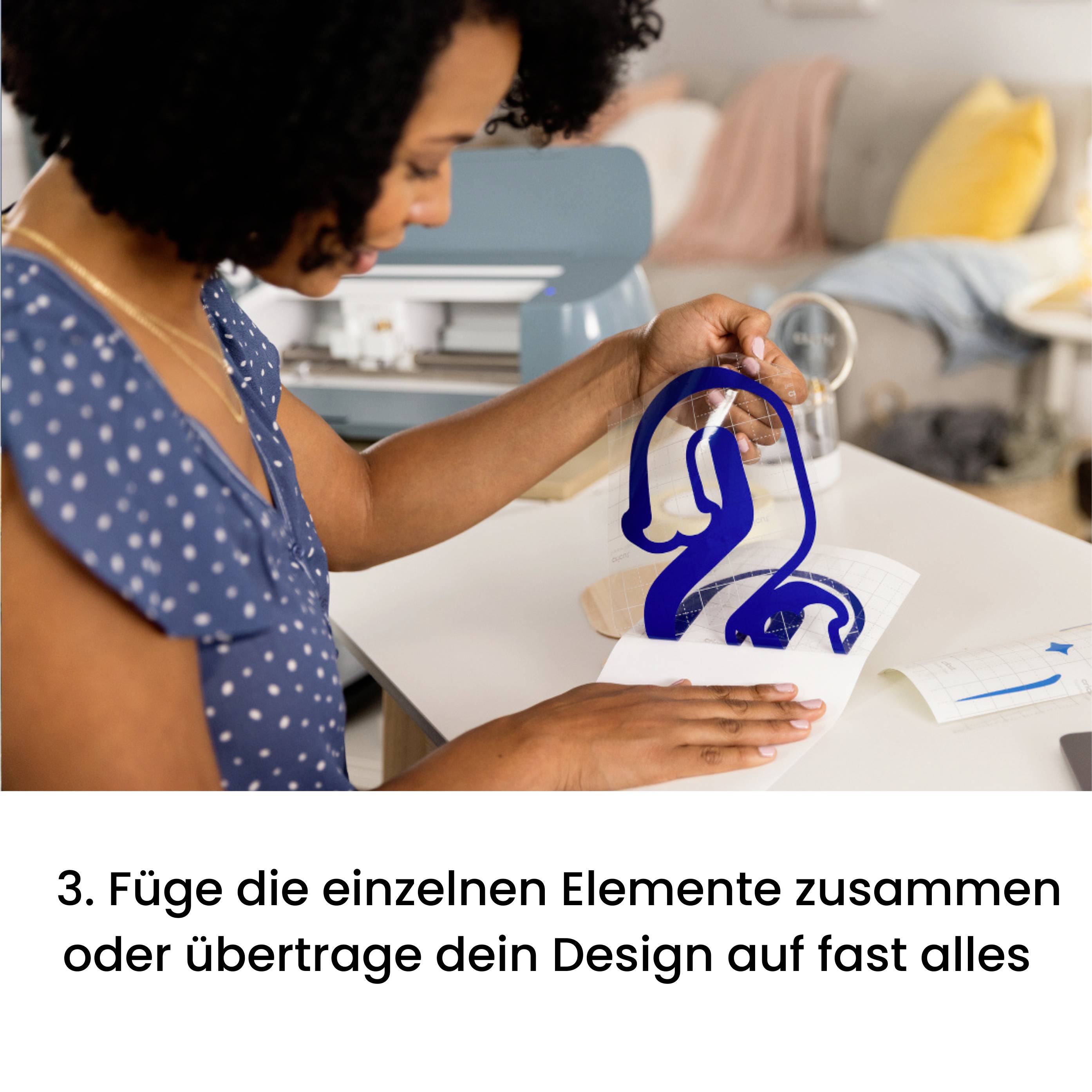 A person is applying blue design elements to an ironing board on a table. A living room is visible in the background.