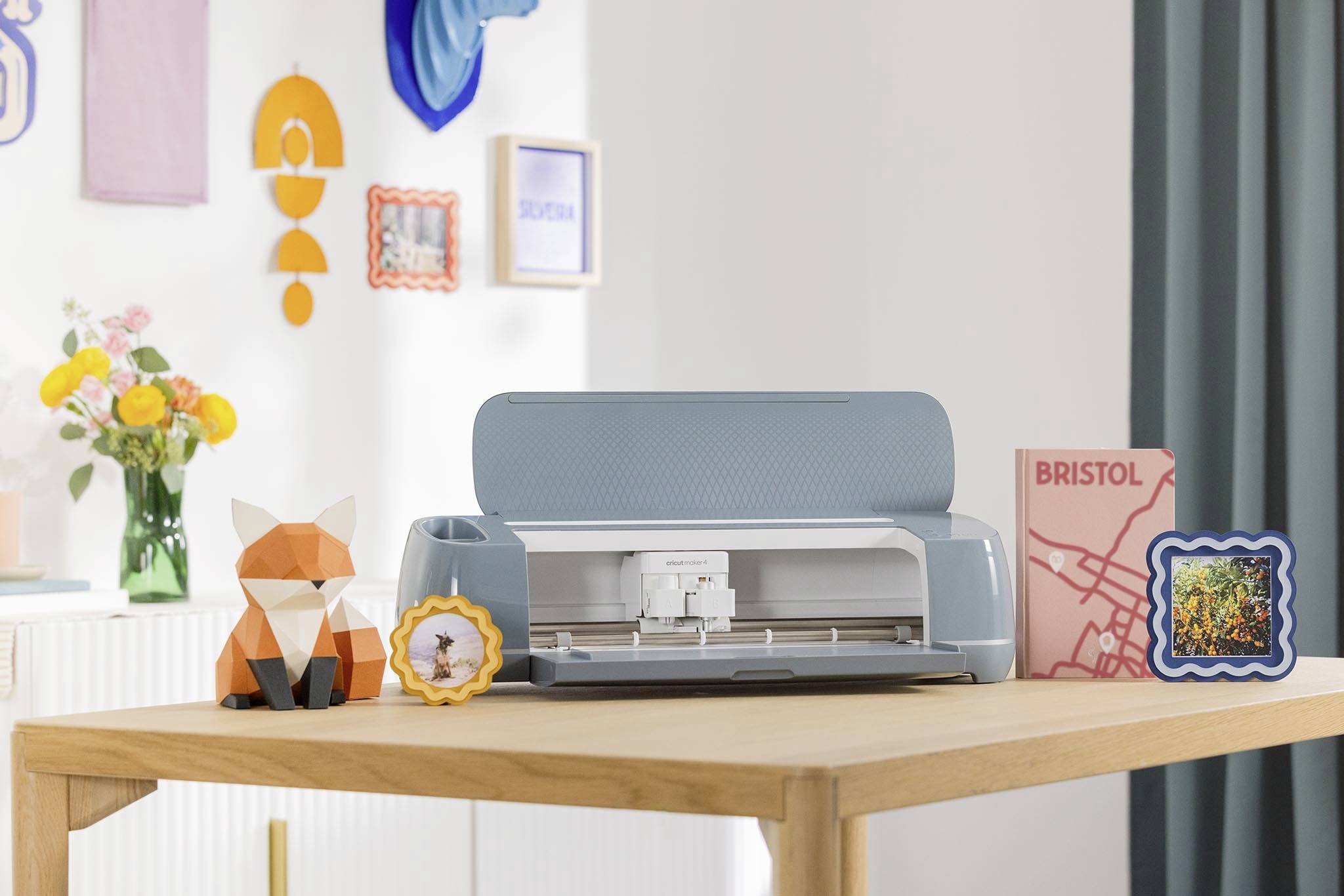 A blue cutting plotter sits on a wooden table, surrounded by colourful decorative items and a floral bouquet in the bright room.