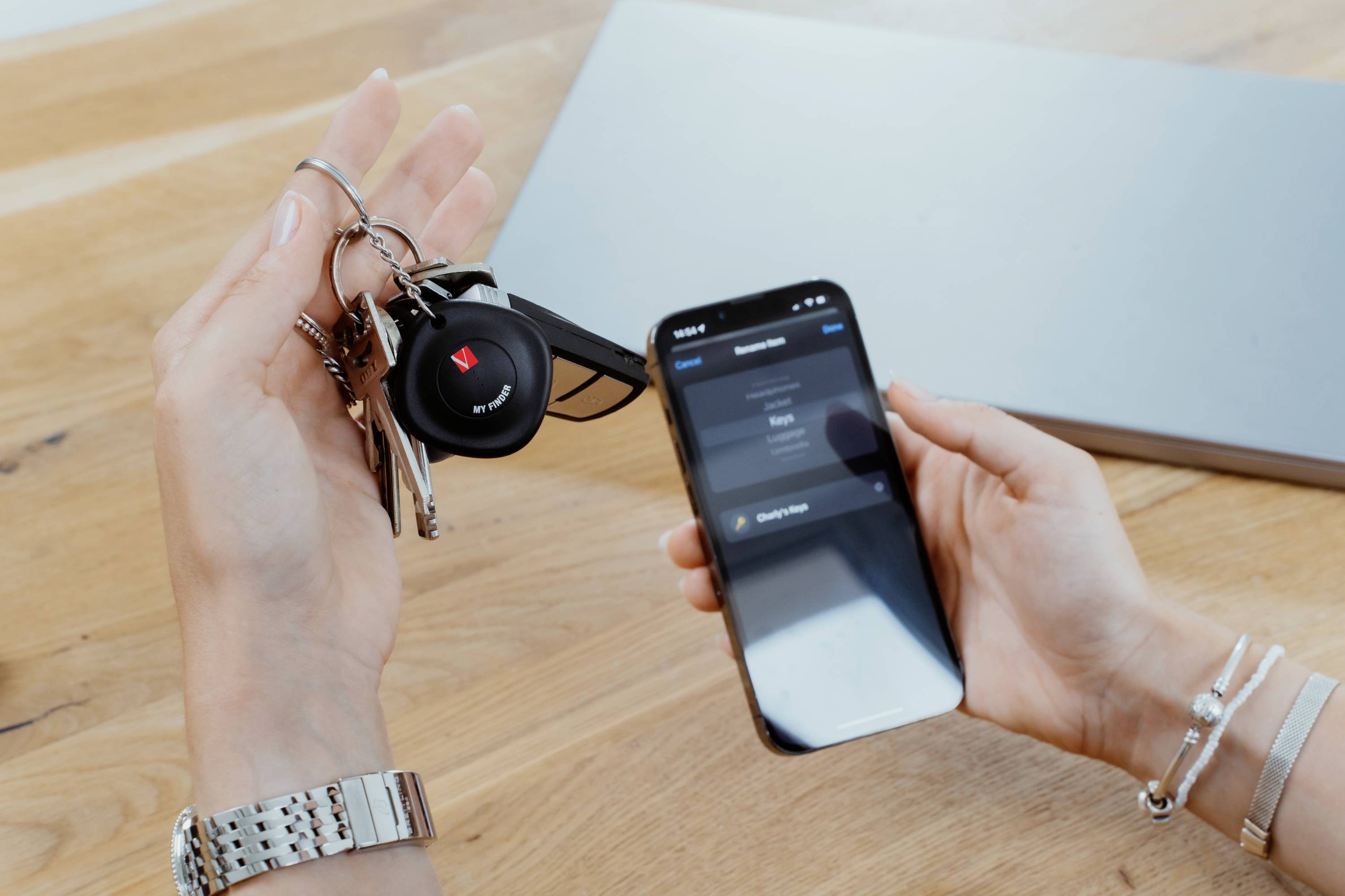 A person is holding a bunch of keys and looking at a smartphone displaying an app. Background: wooden surface, laptop.