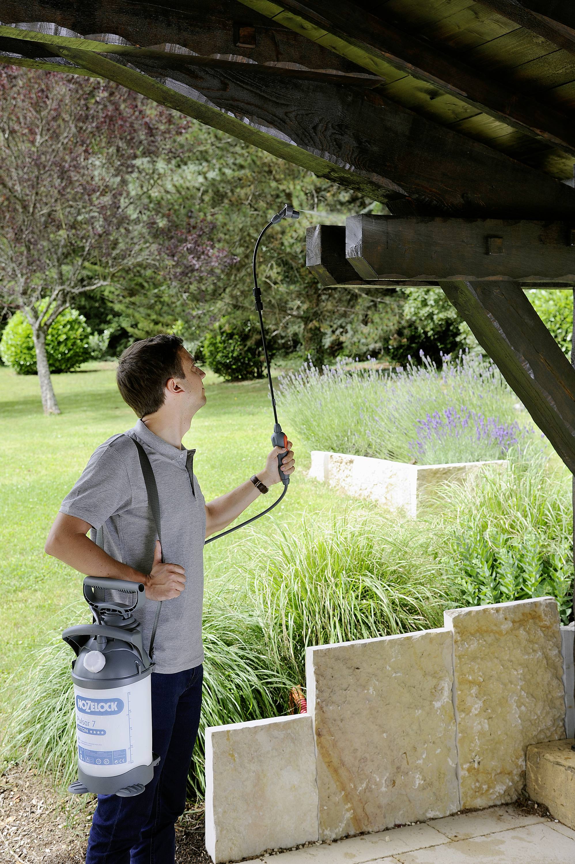 A man is spraying wooden beams of a covered outdoor area in a garden, presumably for protection or cleaning purposes.