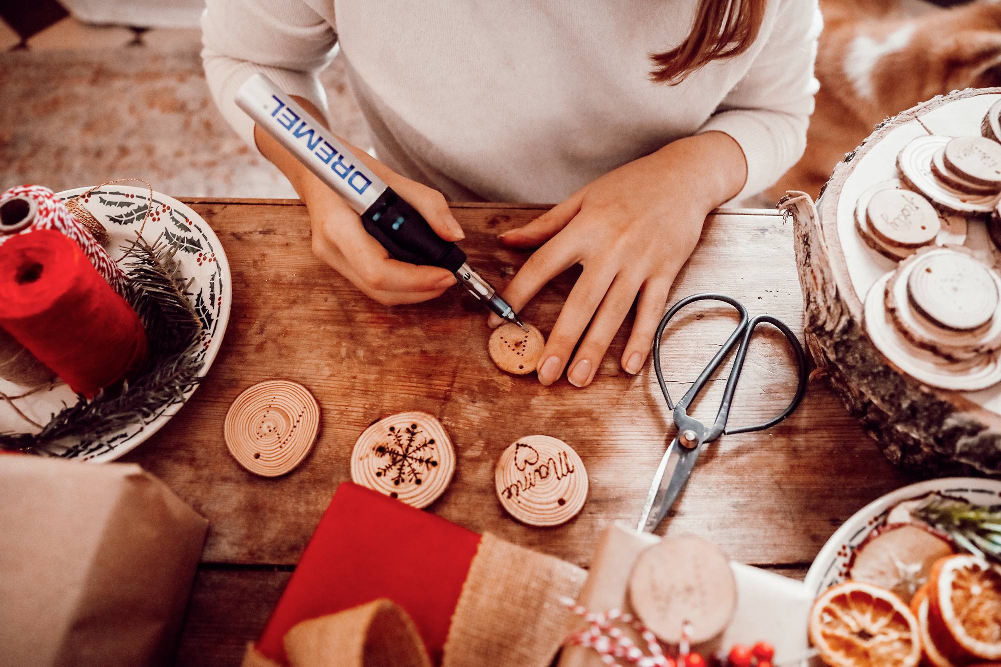 A person is decorating wooden discs with a pyrography pen. Craft materials, scissors, and finished decorations are lying on the table.