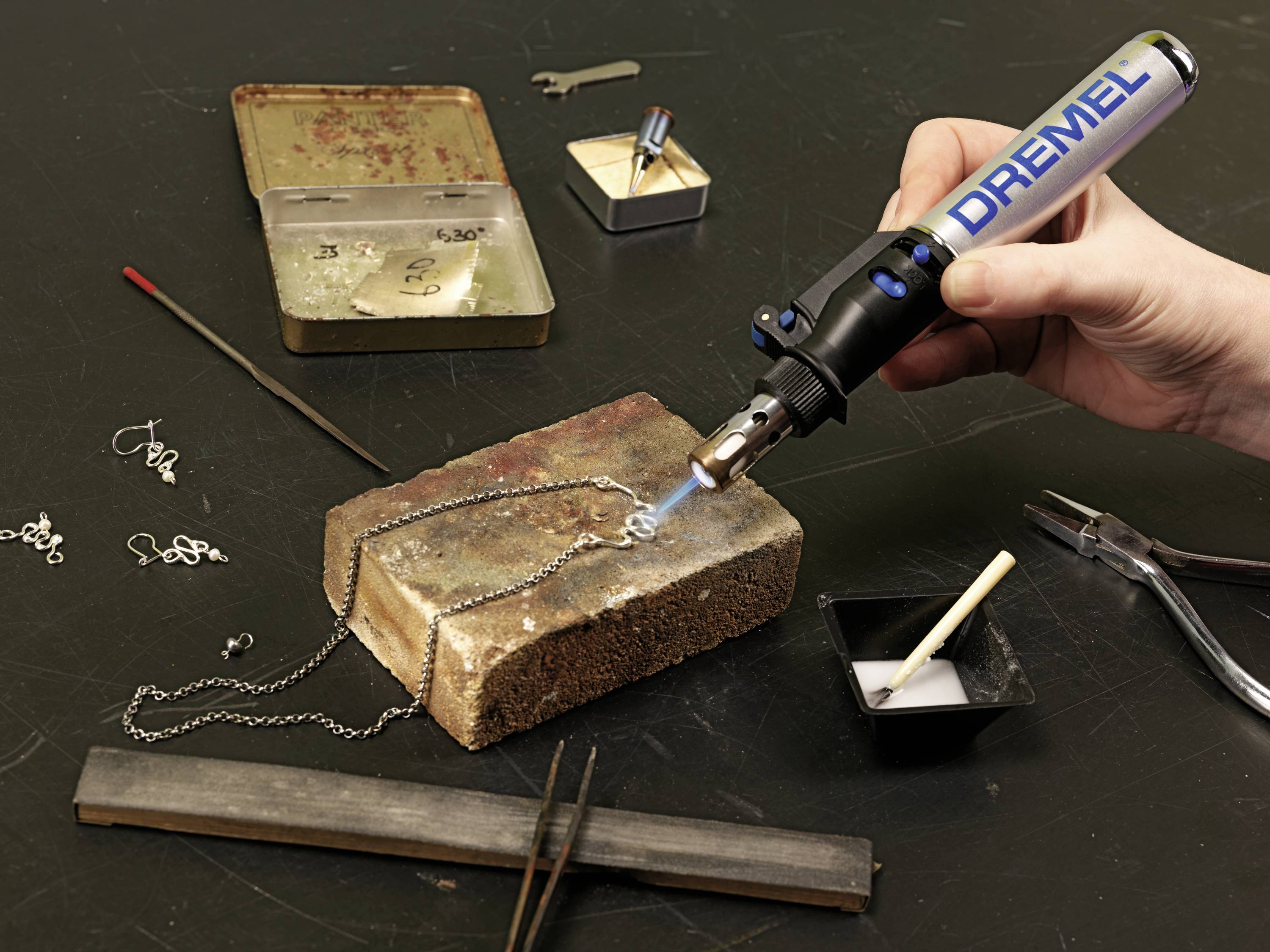 A person is soldering a piece of jewellery onto a stone block using a blow torch. Accessories such as chains, pliers, and flux are visible.