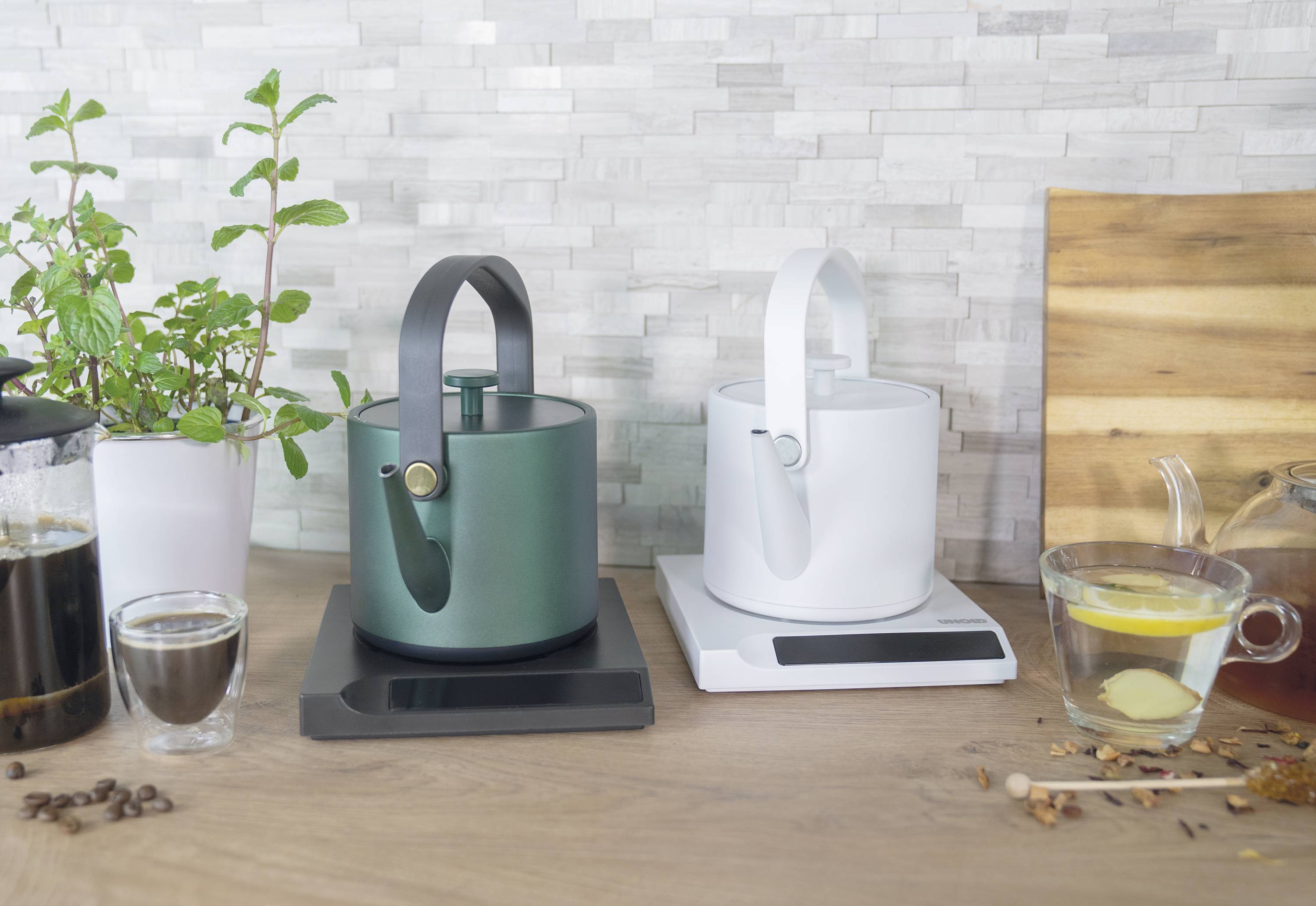 Two modern kettles, one in green and one in white, stand side by side on a countertop, surrounded by plants and beverages.