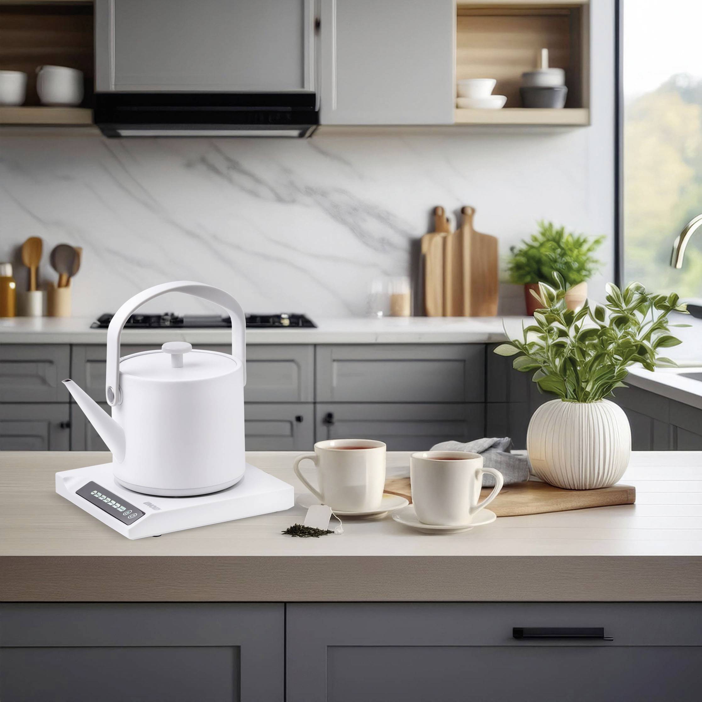 A white electric kettle on a charging station on a kitchen counter, with two cups and a plant next to it; modern kitchen furnishings in the background.