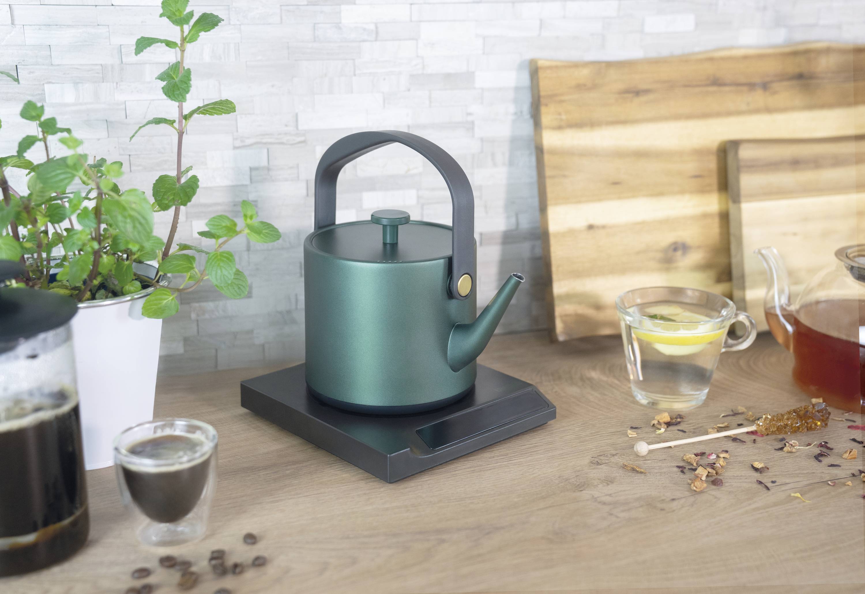 A green kettle sits on a wooden board next to a glass of lemon water, honey sticks, and plants in the background.