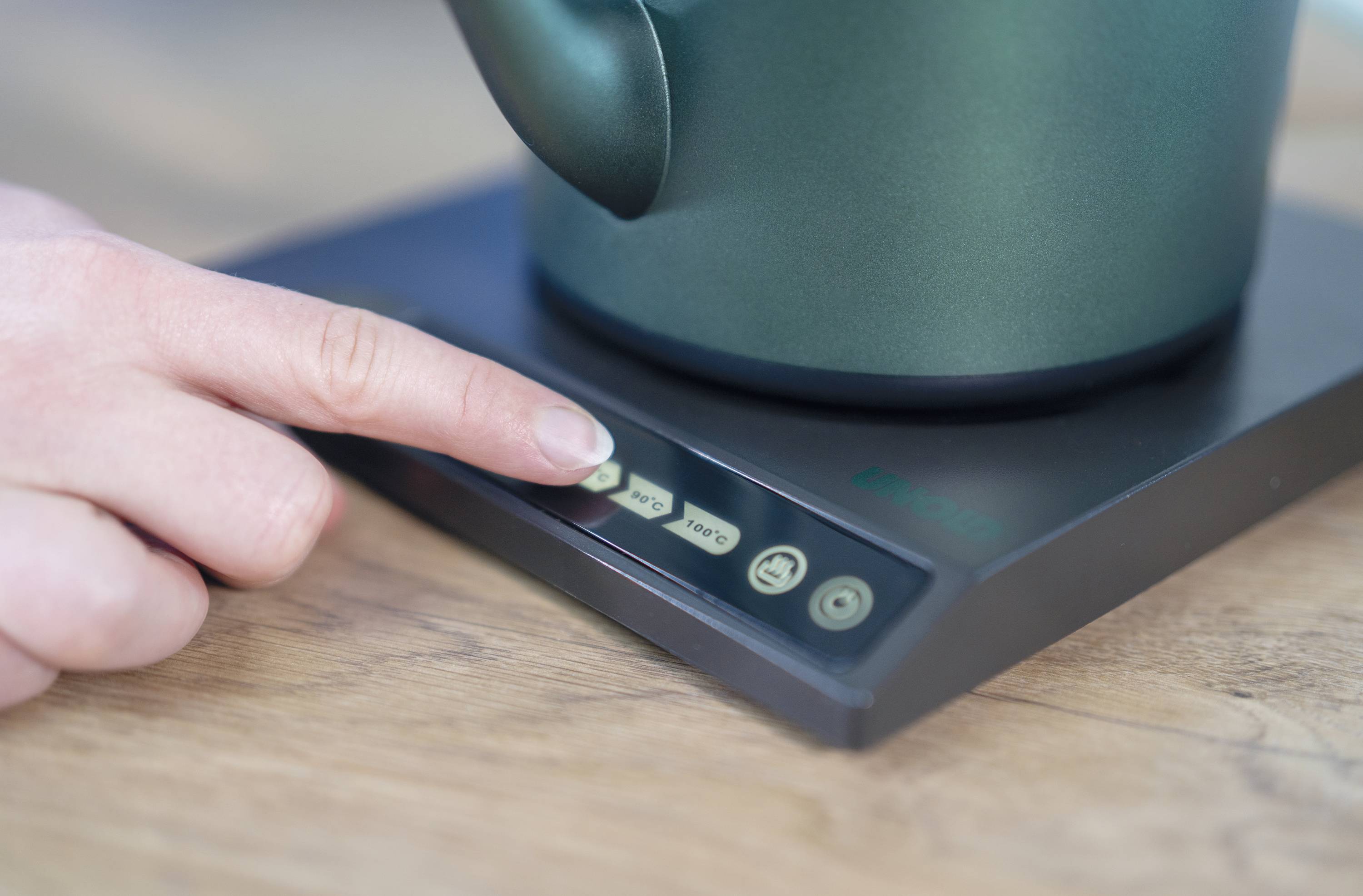 A hand presses a button on an electric kettle. The kettle stands on a wooden surface.