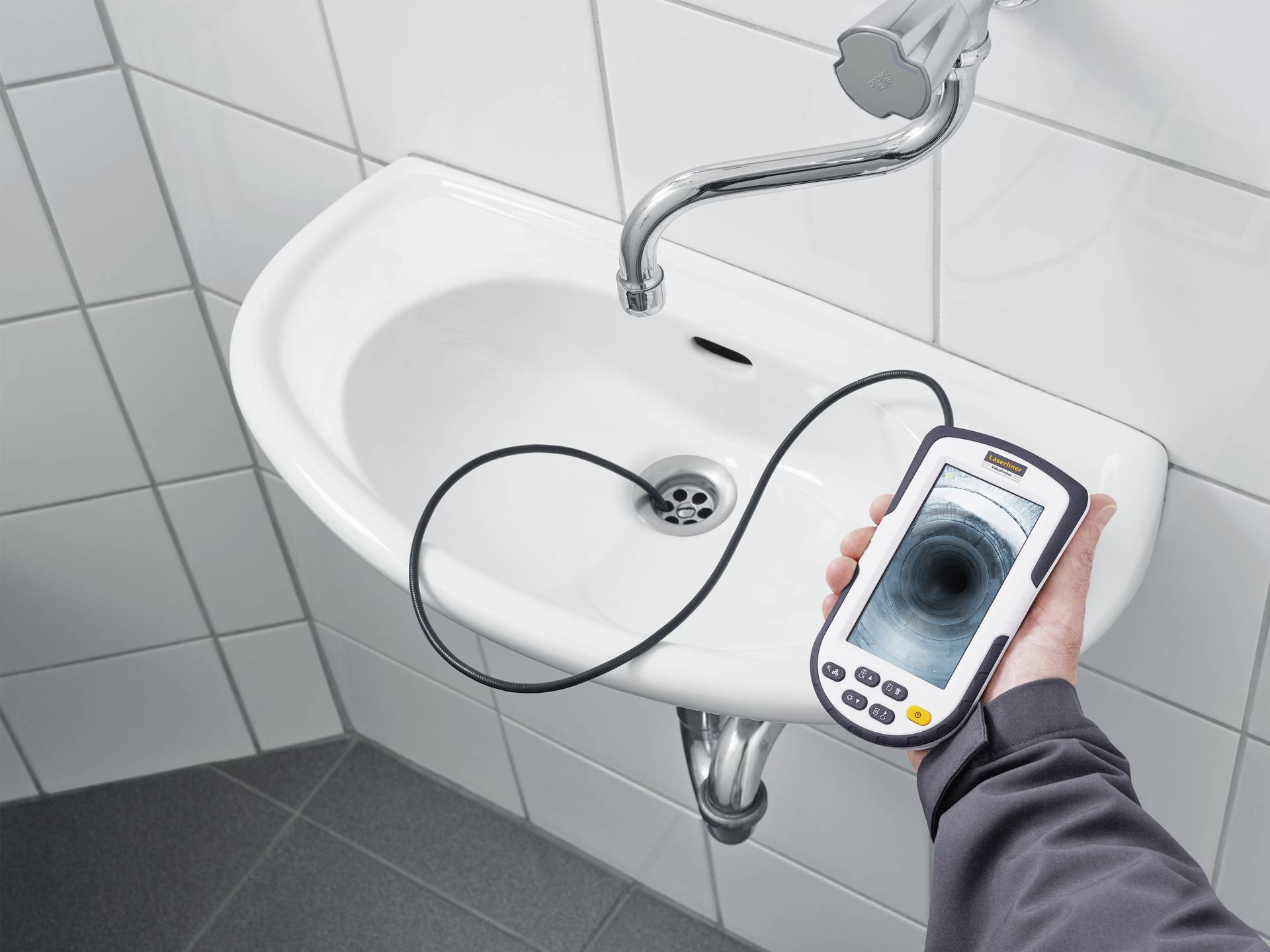 A person is holding an endoscope with a monitor over a sink. The drain pipe is visible on the screen to check for blockages.