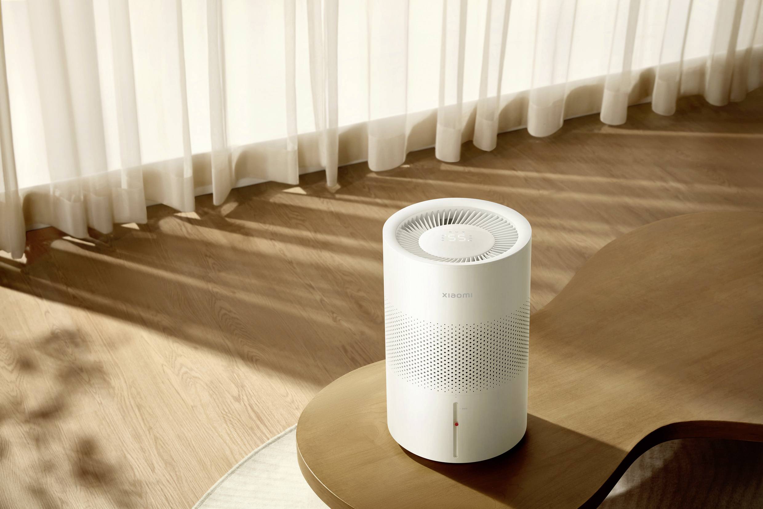 A white, cylindrical air purifier on a curved wooden table. Sunlight filters through curtains, casting shadow patterns.