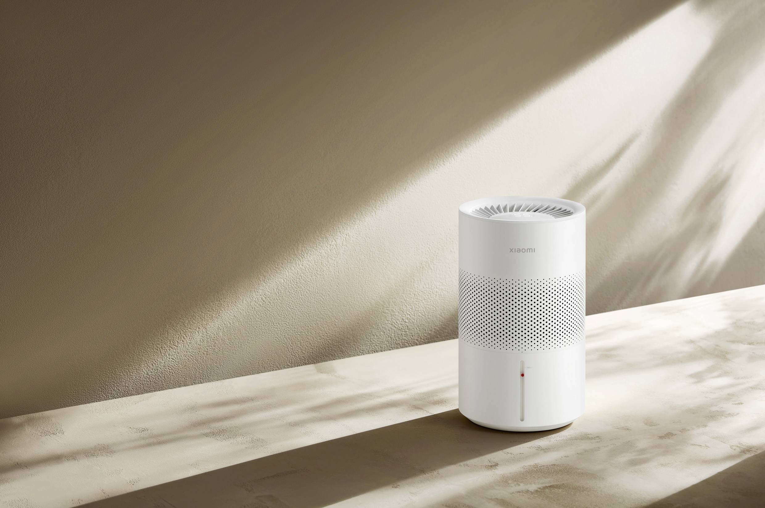 A modern, white air purifier sits on a table surface. Sunlight falls from the side, casting a shadow on the wall.