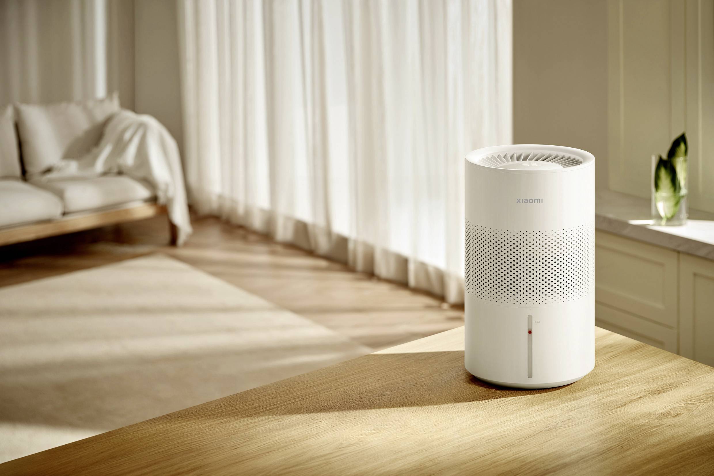 A white air purifier is sitting on a wooden table in a sunlit room with a sofa and light-coloured curtains in the background.