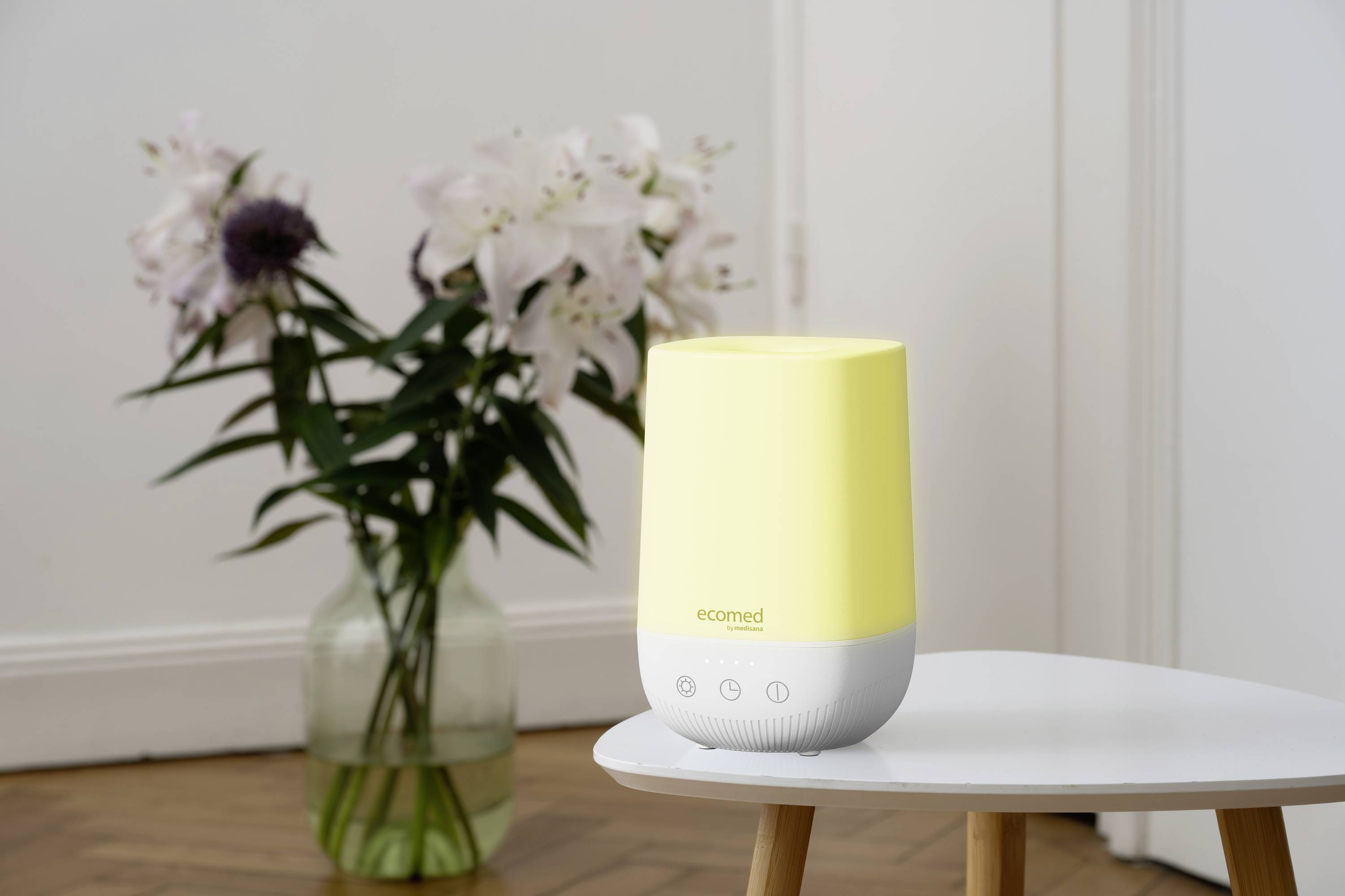 A yellow humidifier on a table in front of a flower bouquet in a vase, captured in a bright room with a wooden floor.