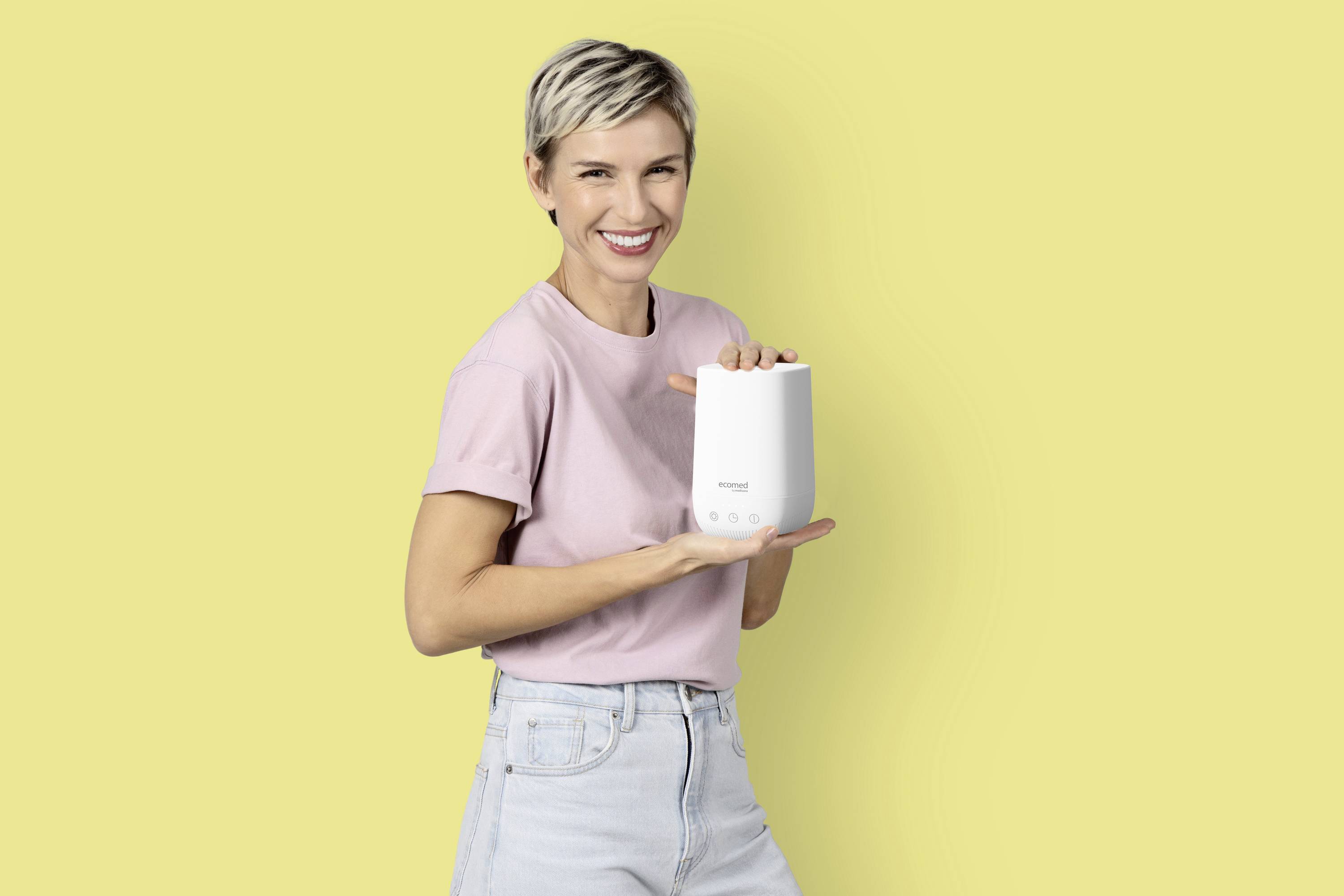 A woman smiles while holding a router against a yellow background. She is wearing a pink T-shirt and jeans, presenting the device with its recognisable logo and control buttons.