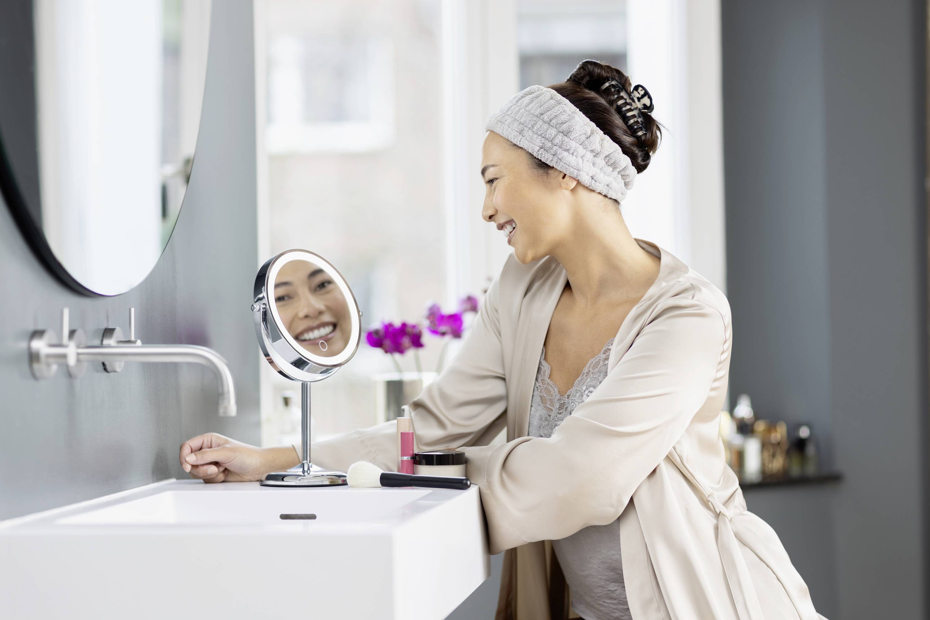 A woman smiles at herself in the mirror in a modern bathroom. She is wearing a dressing gown and a headband. Cosmetic items are placed on the washbasin.