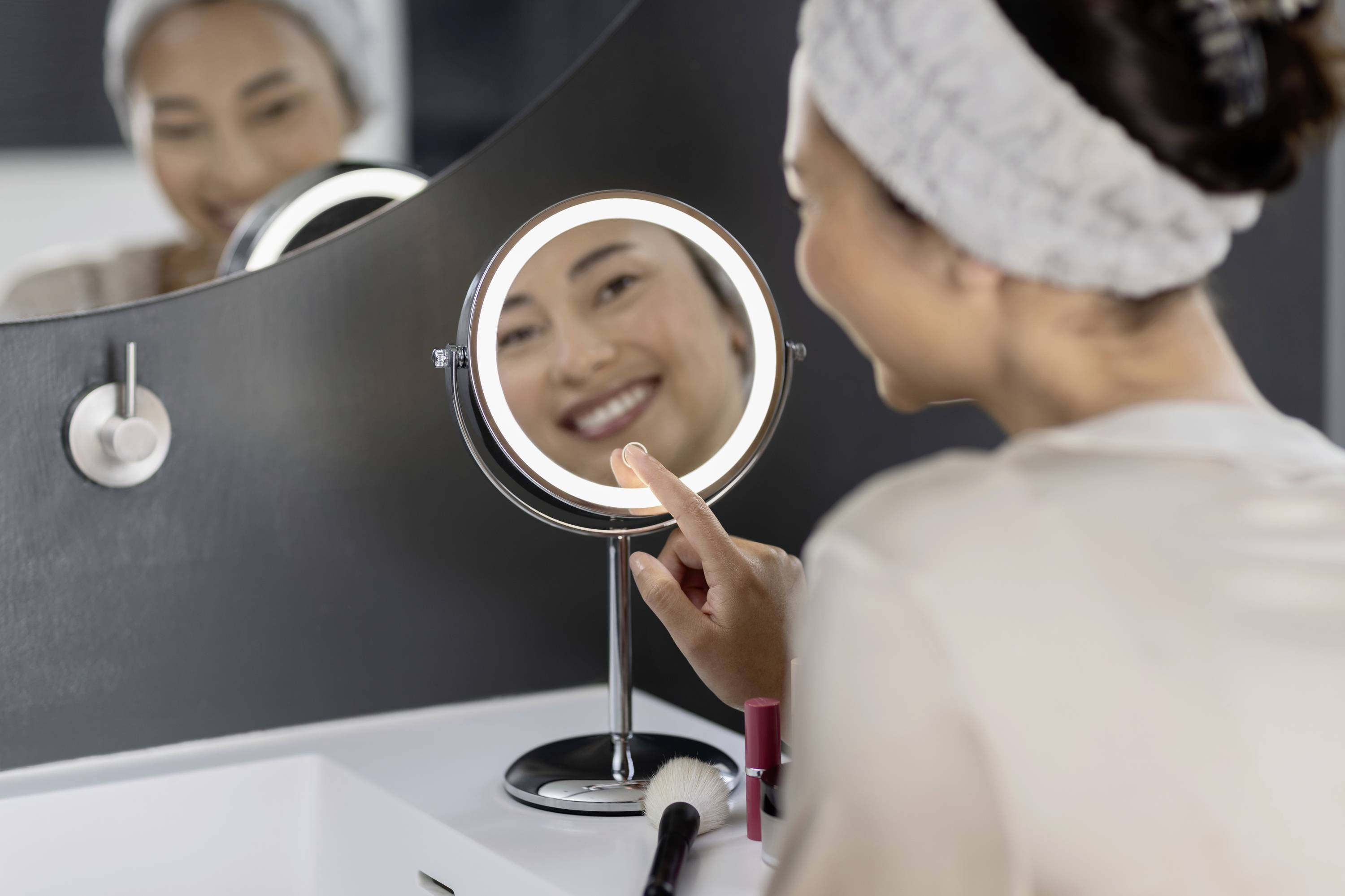 A person touches an illuminated make-up mirror while smiling into it. In the foreground is a sink with cosmetic accessories.