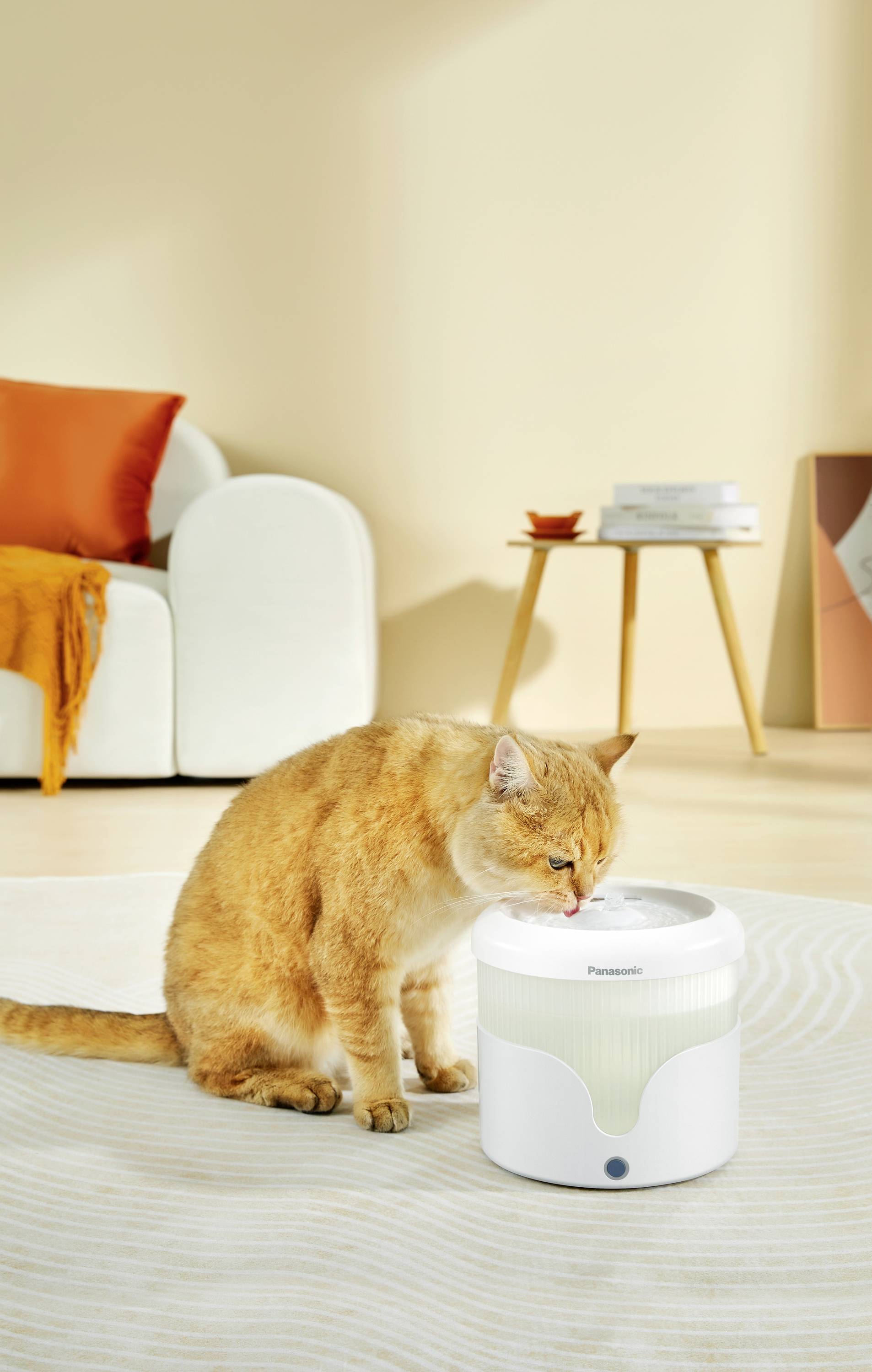 An orange cat is drinking from a white water dispenser in a bright living room with a sofa and a side table in the background.