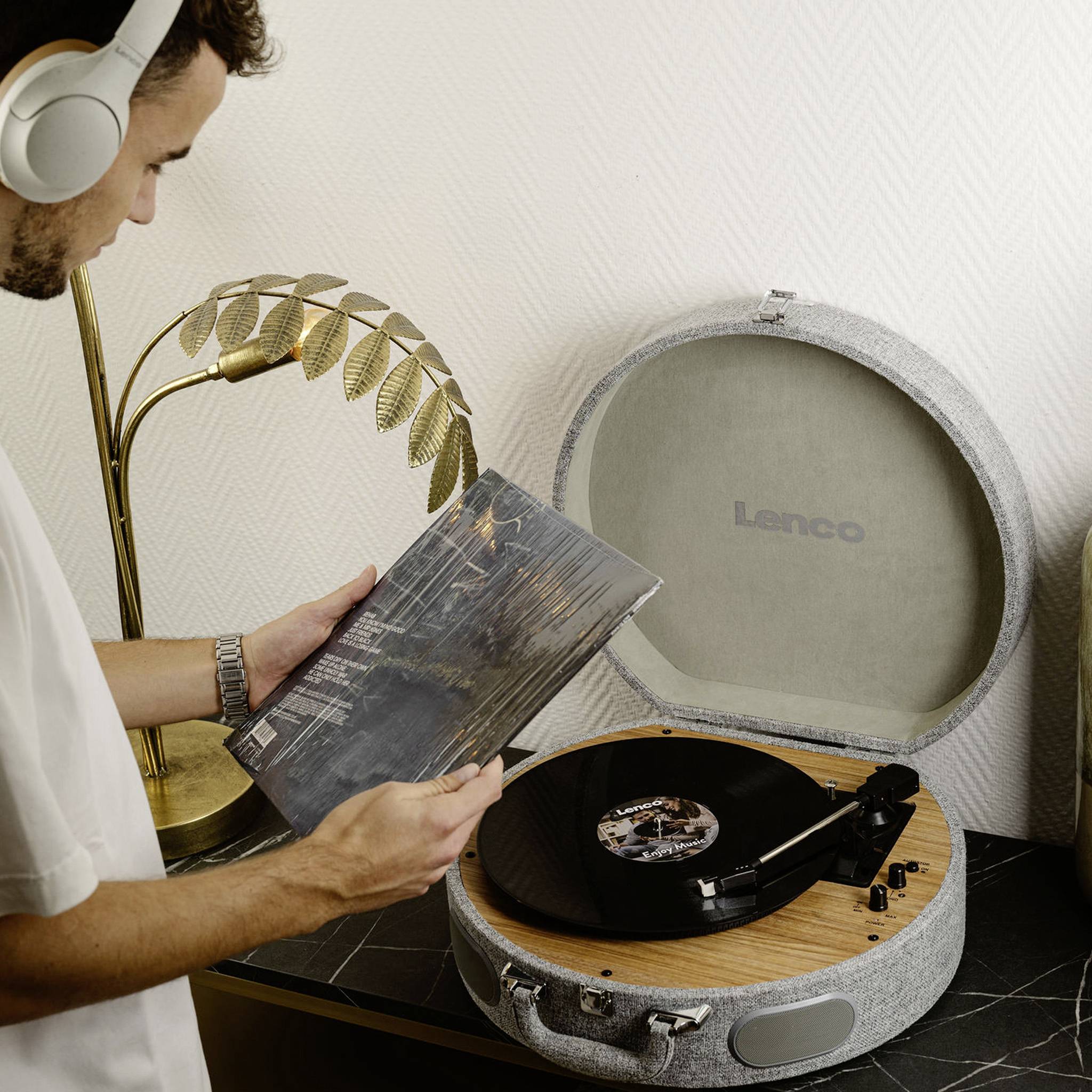 A person holds a vinyl record in front of a record player while wearing headphones. The background features a lamp and a white wall.