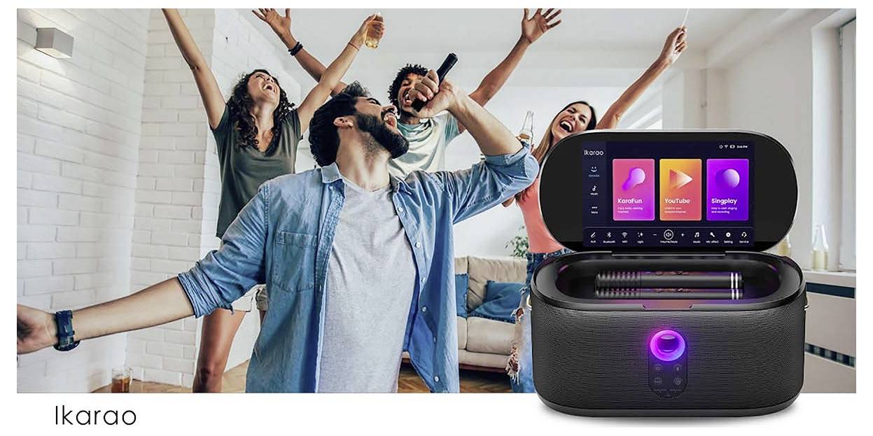 Group of people singing and dancing in a living room, next to a karaoke machine with a screen displaying song selections and controls.