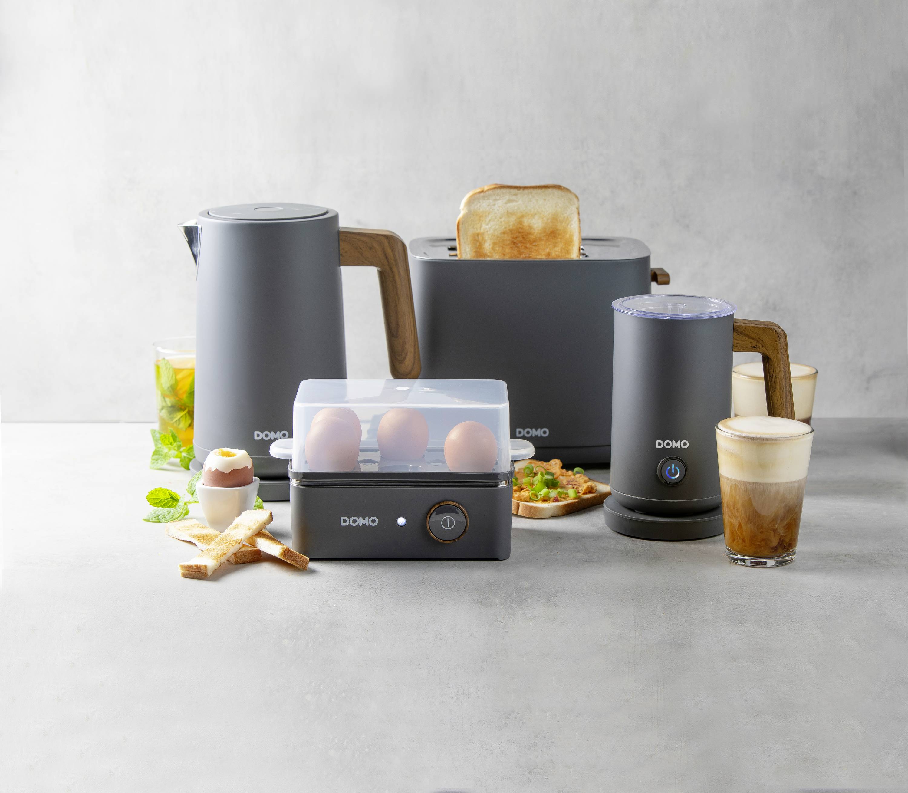 Several kitchen appliances, including a toaster with toast, an electric kettle, an egg cooker with eggs, and a milk frother beside drinks.