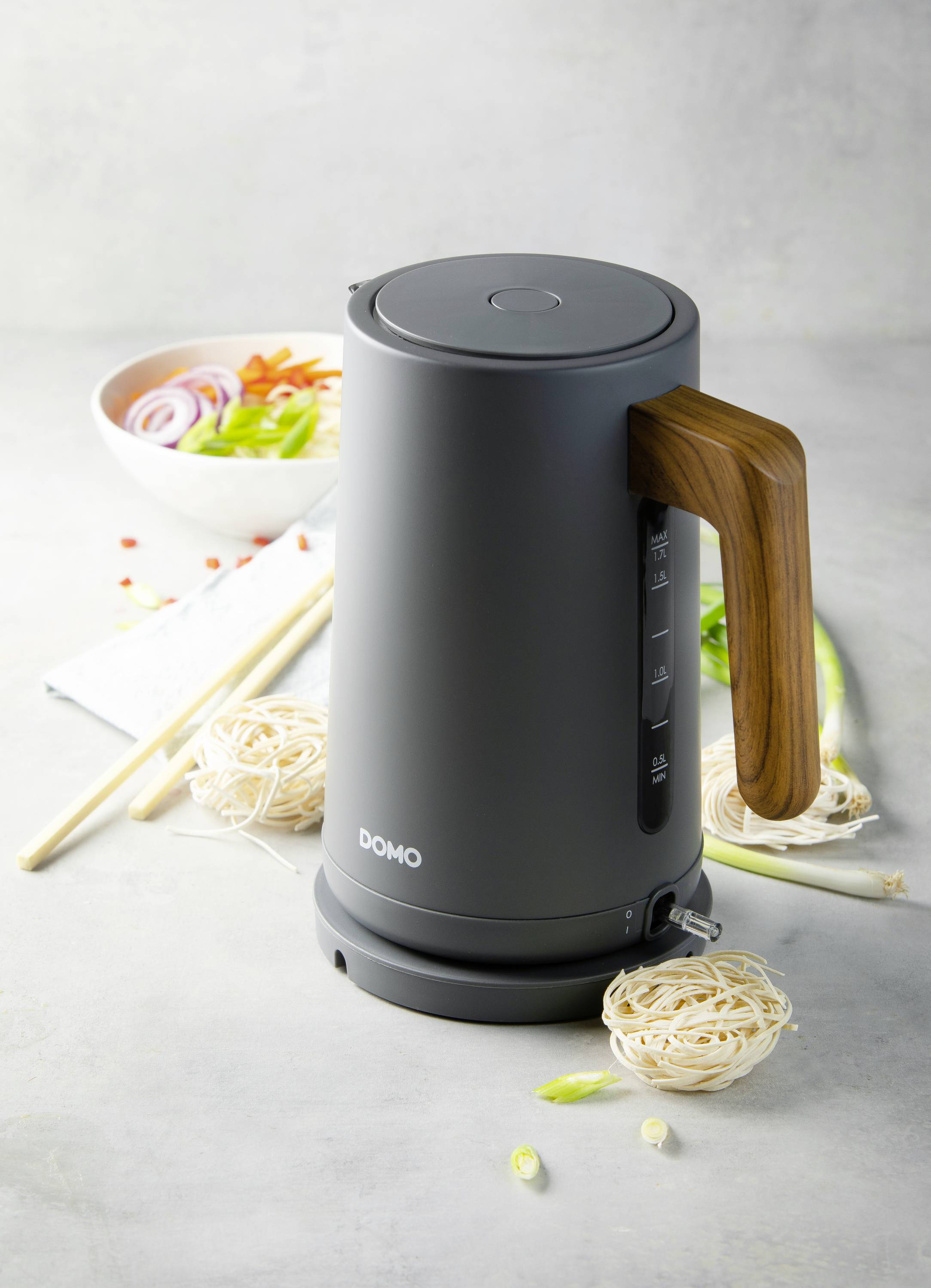A black kettle with a wooden handle sits on a table, surrounded by pasta and vegetables. In the background, a bowl of chopped vegetables.
