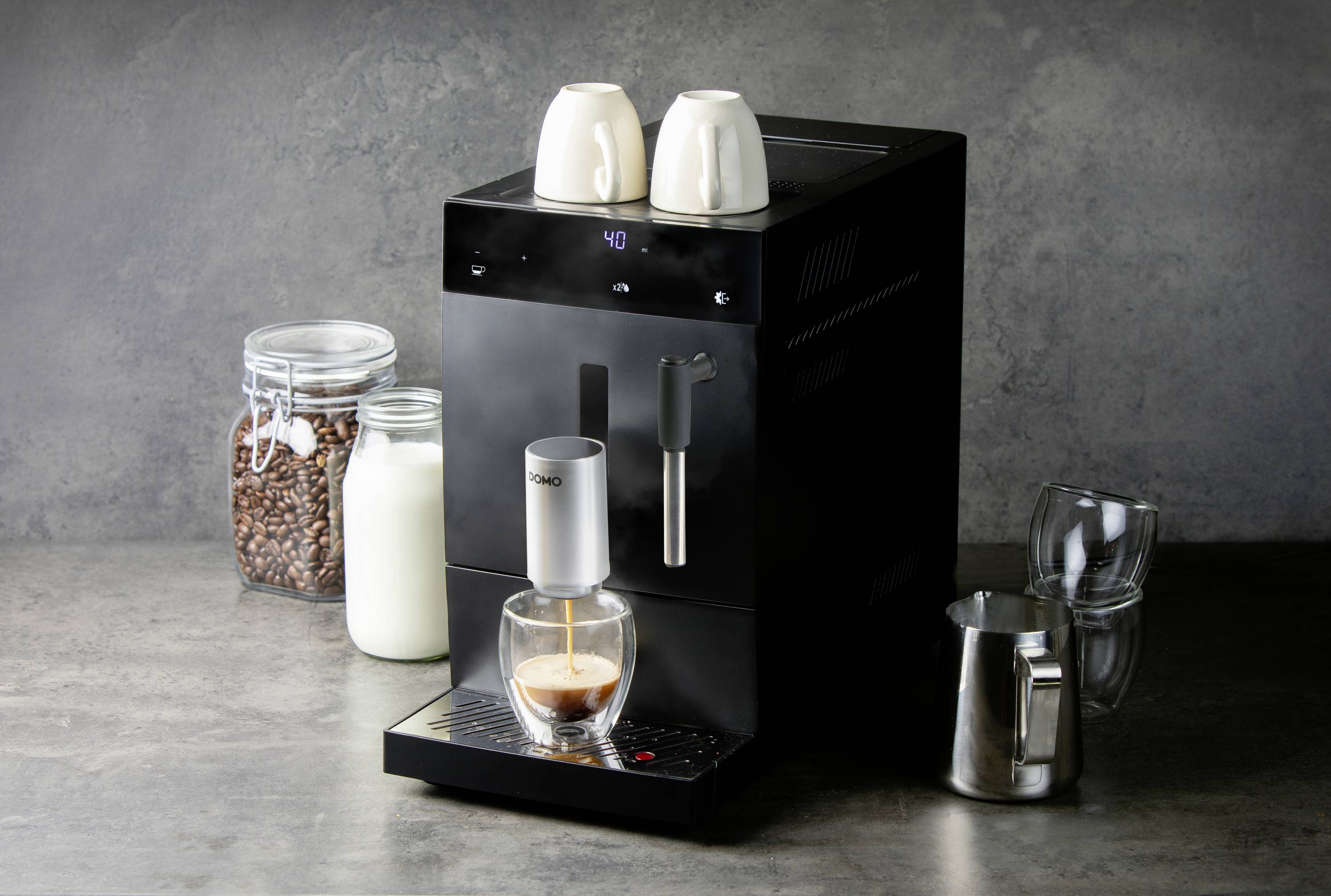 Black coffee machine on a grey background, with coffee cups on top, milk, beans, and glasses beside it. Coffee is being brewed into a glass.