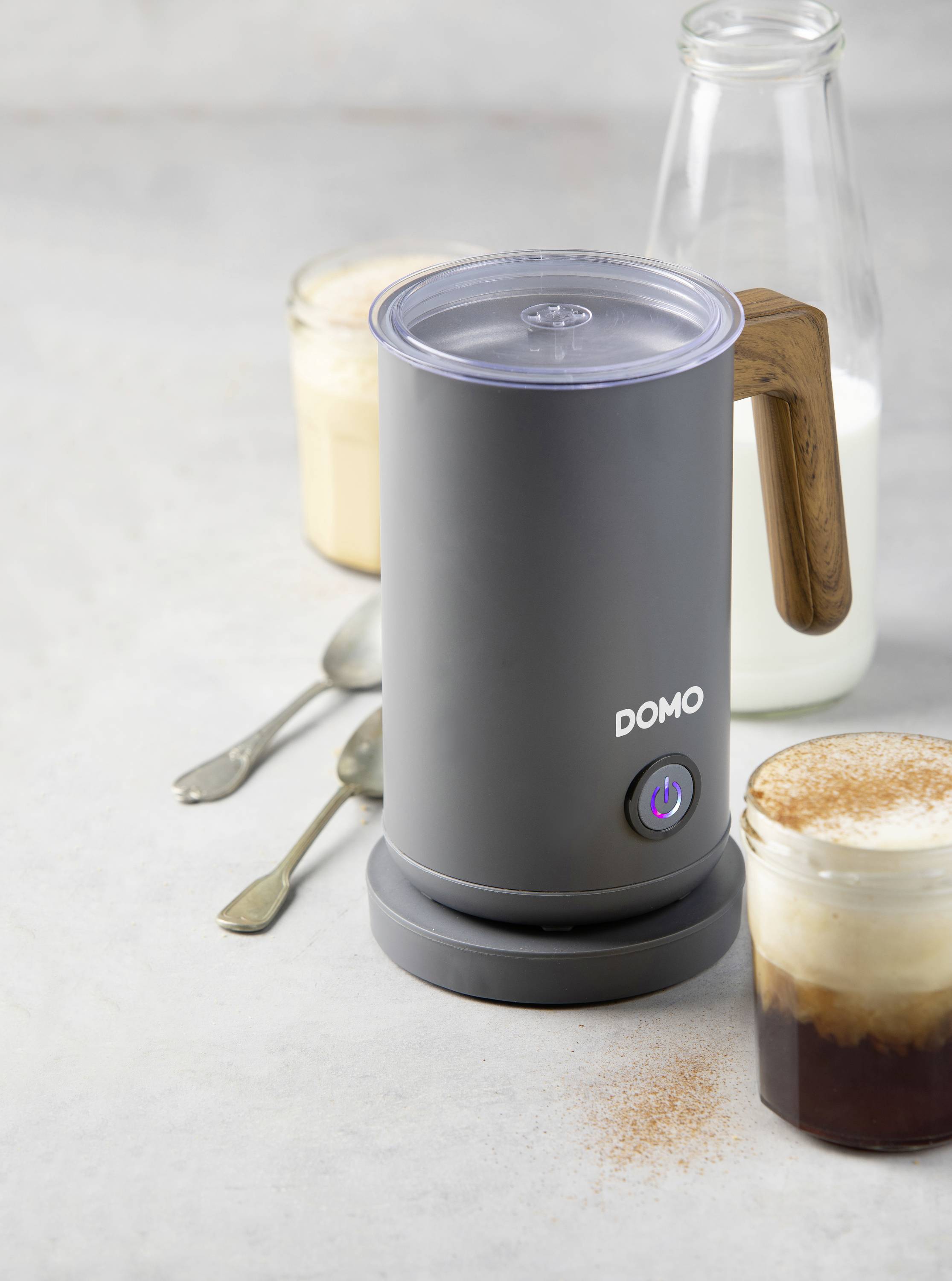 An electric milk frother with a wooden handle sits on a table next to two glasses of café latte and a glass of milk.