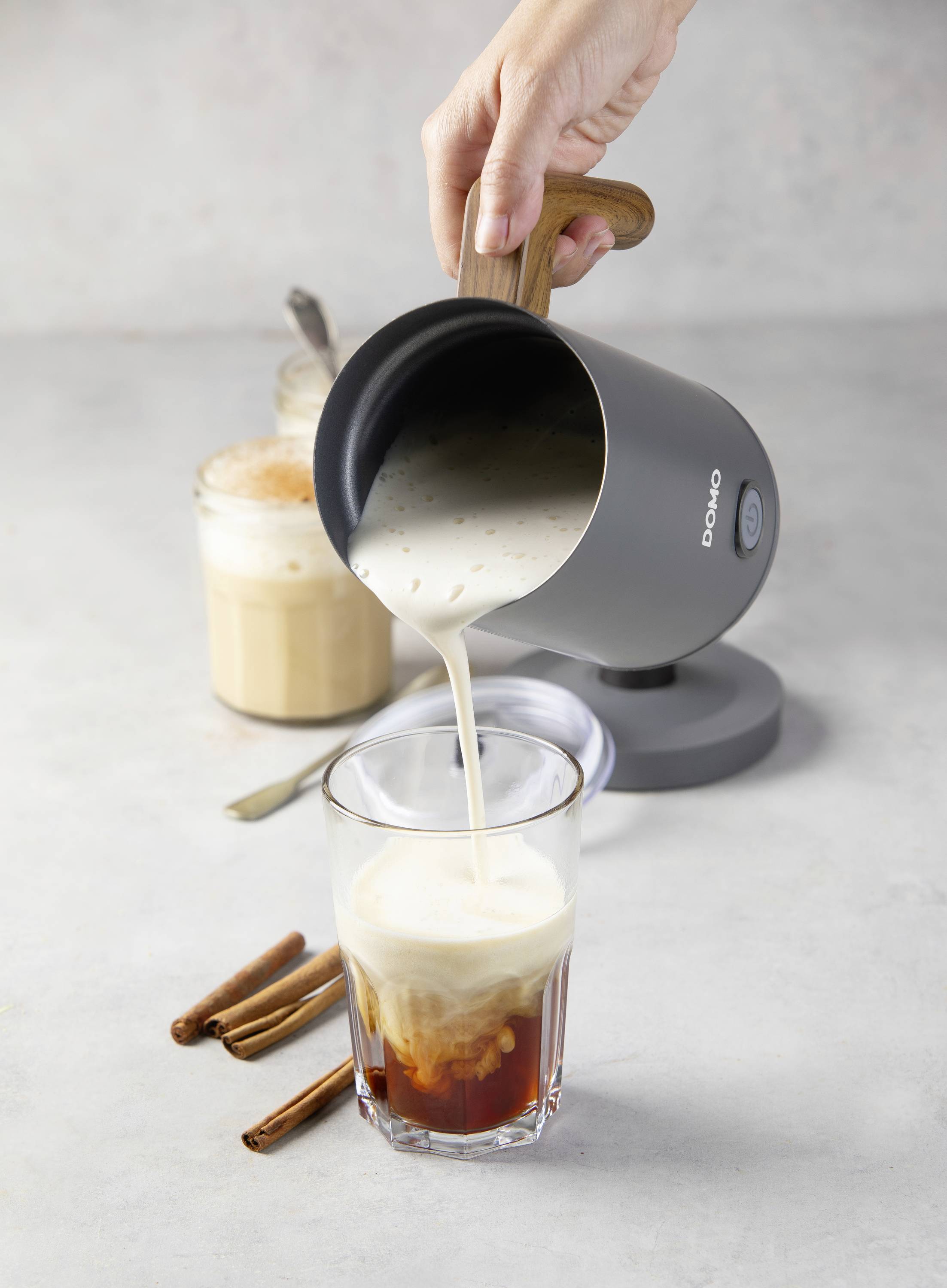 A hand is pouring milk from an electric milk frother into a glass, with coffee and cinnamon sticks beside it on a table.