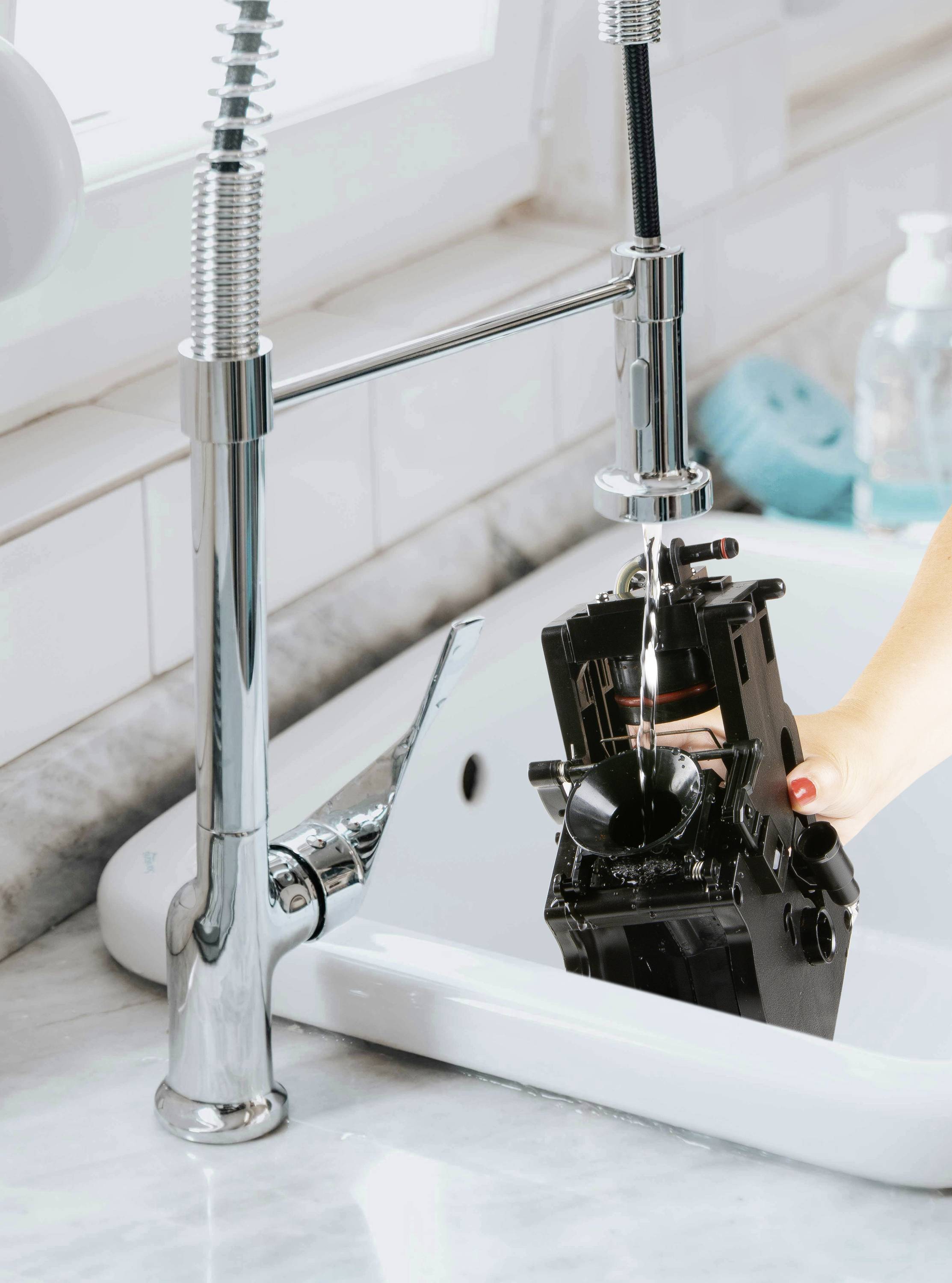 A hand is washing a camera under a tap in a sink. The scene takes place in a bright kitchen with white tiles.