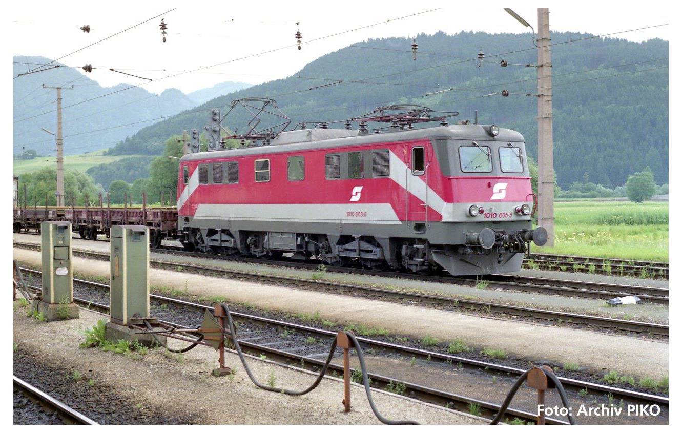 A red locomotive of the Austrian Federal Railways travels along a track in front of a mountainous landscape. Power lines run along the left side.