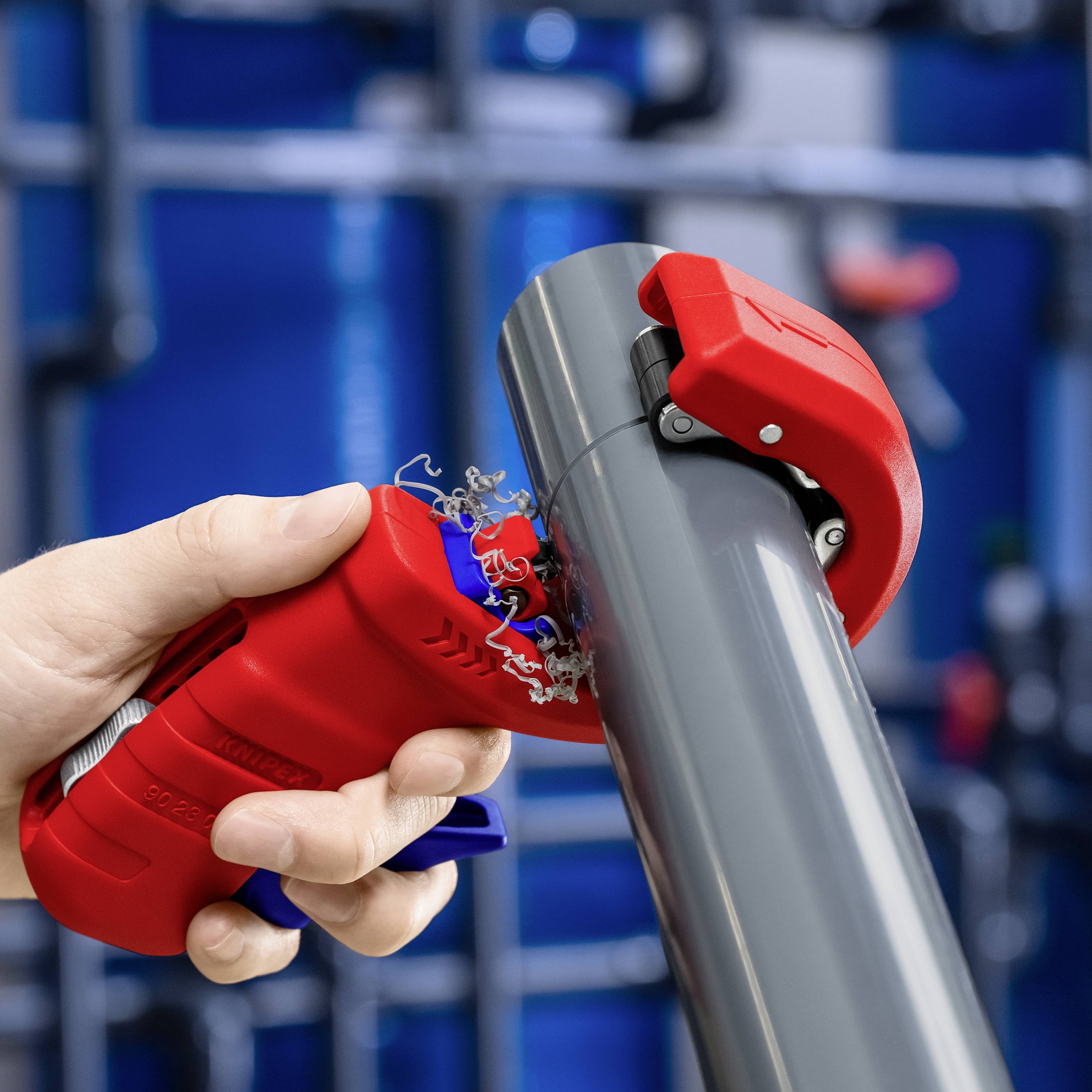 A person is cutting a grey pipe with a red pipe cutter. Blurry industrial pipelines are visible in the background.