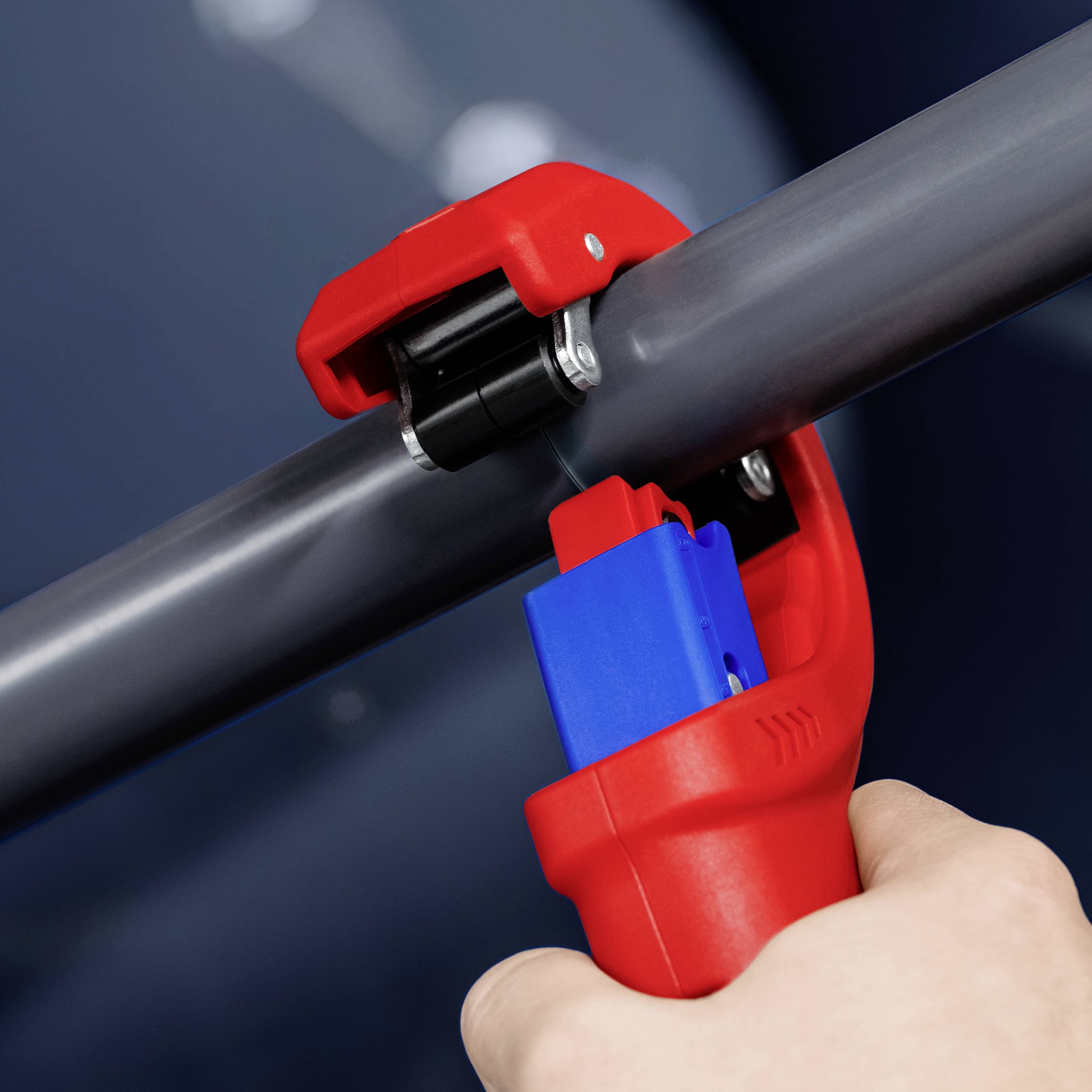 A person is cutting a grey pipe with a red pipe cutter. Focus on the tool.