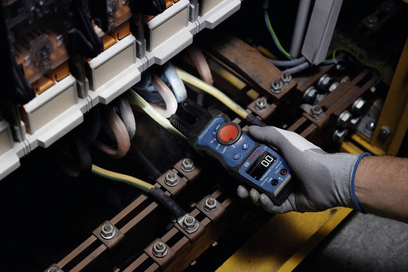 A person is measuring the voltage in an electrical control cabinet using a digital multimeter. Gloves protect the hands.