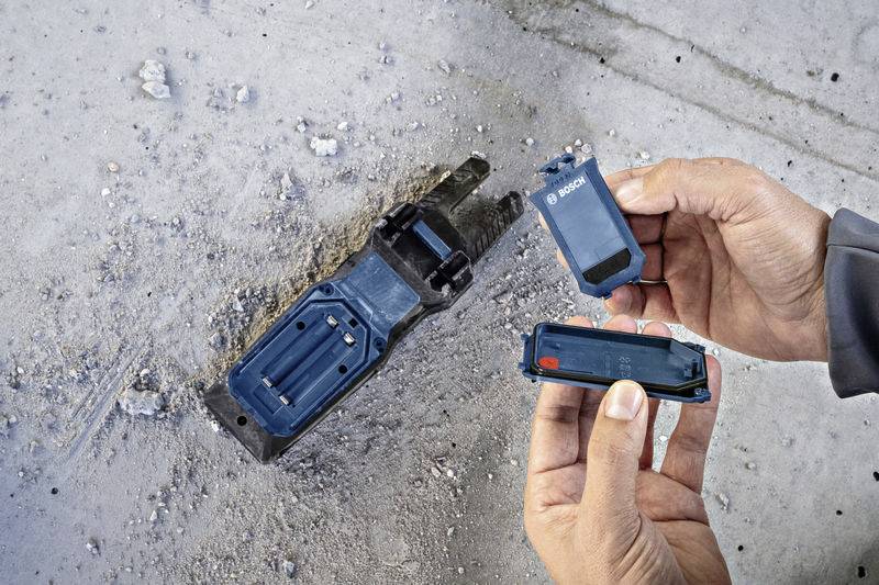 Two hands are holding a Bosch screwdriver attachment and a battery. Another Bosch tool lies on a concrete-like floor.
