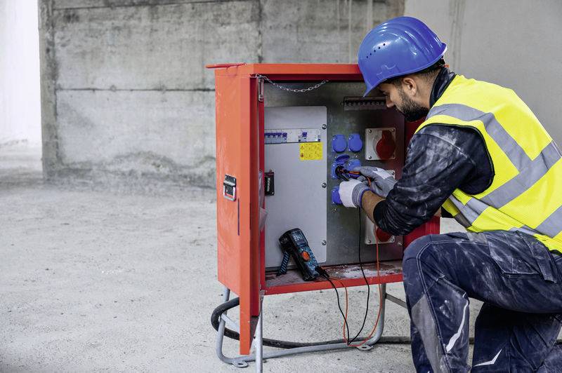A worker wearing a blue hard hat and high-visibility vest is inspecting an electrical control cabinet on a construction site.