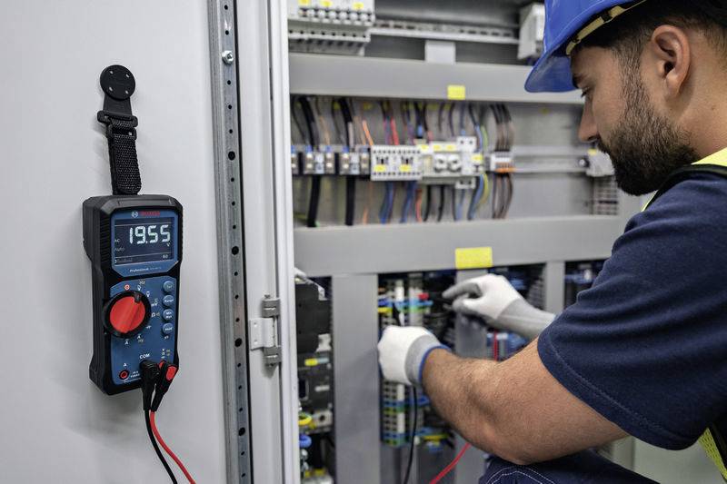 An electrician wearing a safety helmet is working on an electrical cabinet. A multimeter displays '19.55'. He is checking and wiring the installations.