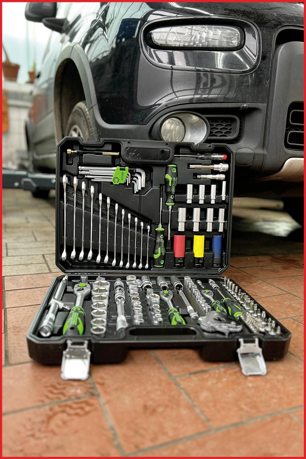 An open toolbox with various spanners, socket wrenches, and bits is lying on the ground in front of a car.