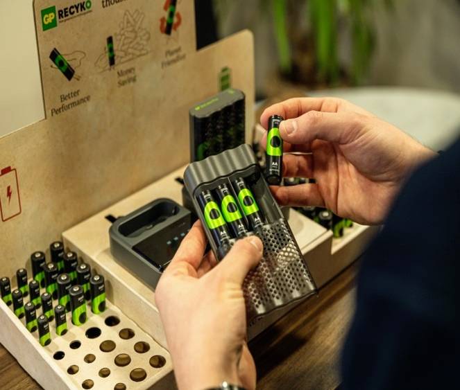 Person hält Batterien eines Ladegeräts an einem Verkaufsstand. Auf Kartonverpackung steht: 'Better Performance', 'Money Saving', 'Planet Friendly'.