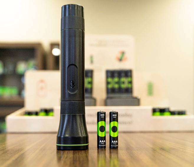 A black torch stands upright on a table, surrounded by two AAA batteries. Blurred shelves are visible in the background.