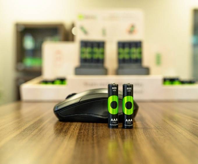 Two green AAA batteries are in the foreground on a table, with a computer mouse beside them. Further batteries can be seen in a display in the background.