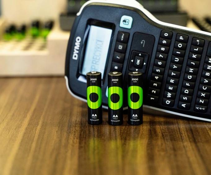 A label printer and three rechargeable AA batteries on a wooden table. The printer displays a screen and a keyboard.