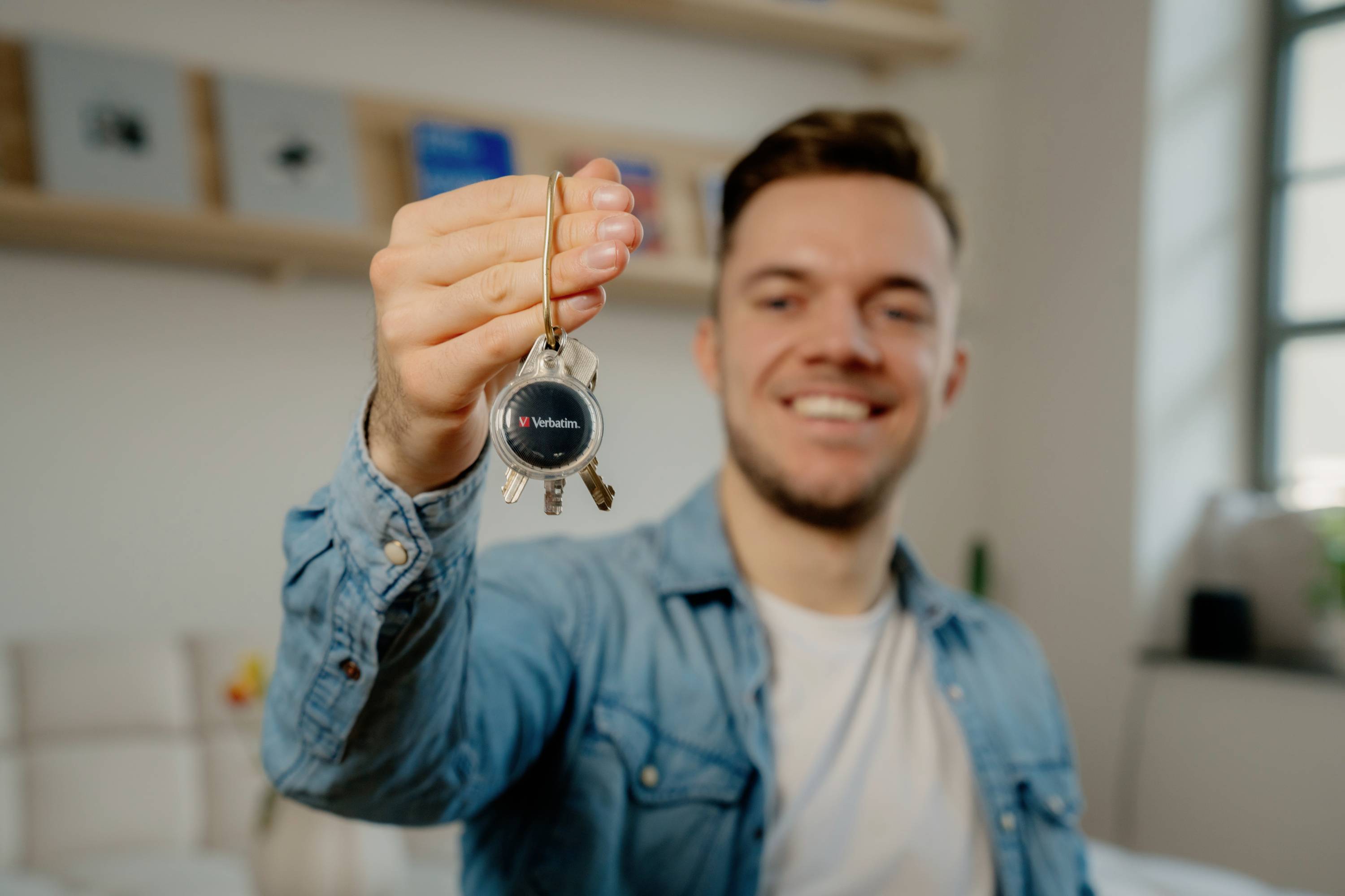 A person holds a bunch of keys in front of the camera, smiles, and is wearing a denim jacket. Shelves are visible in the background.