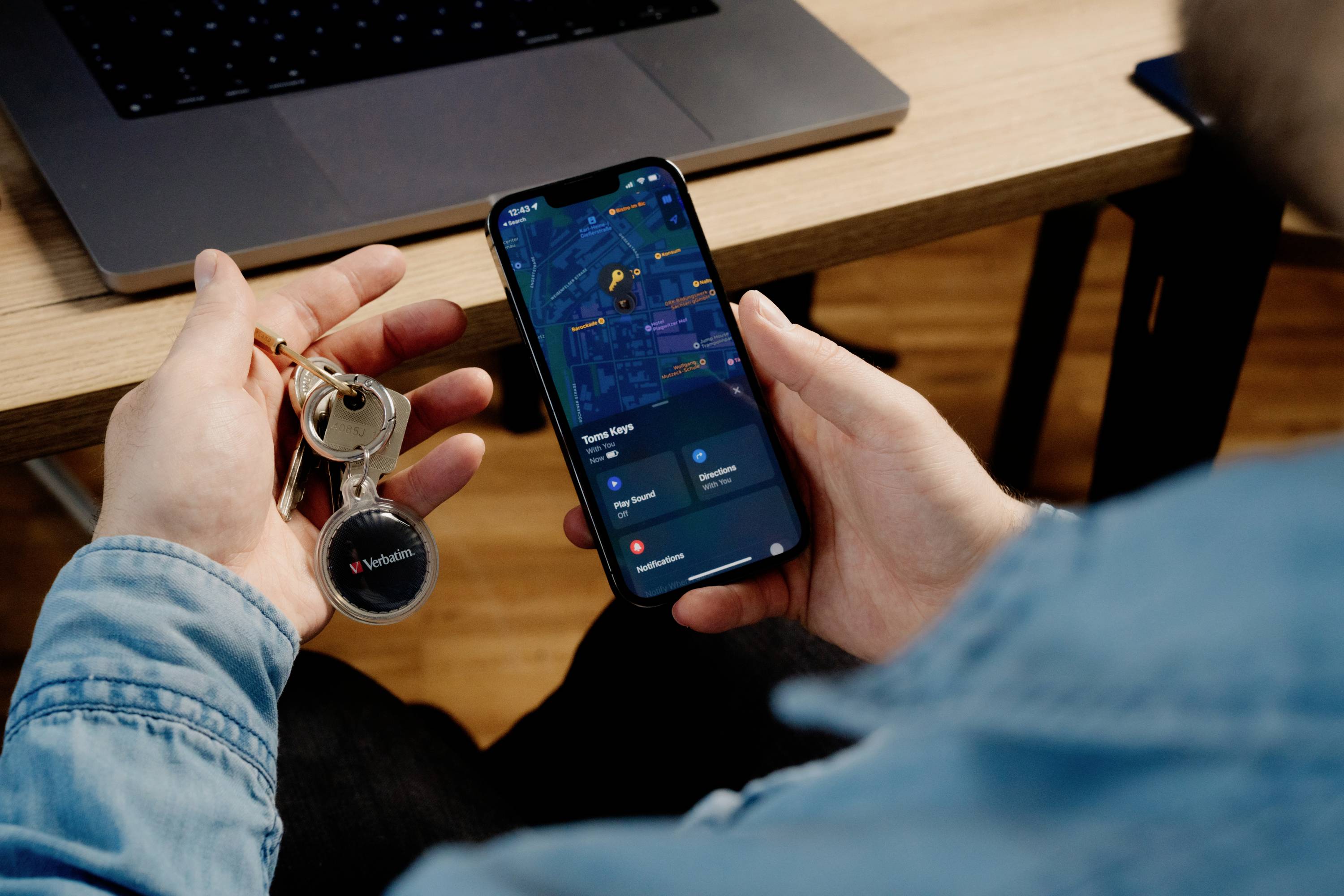 A person is sitting in front of a laptop, holding a smartphone with an open maps app in one hand and keys in the other.