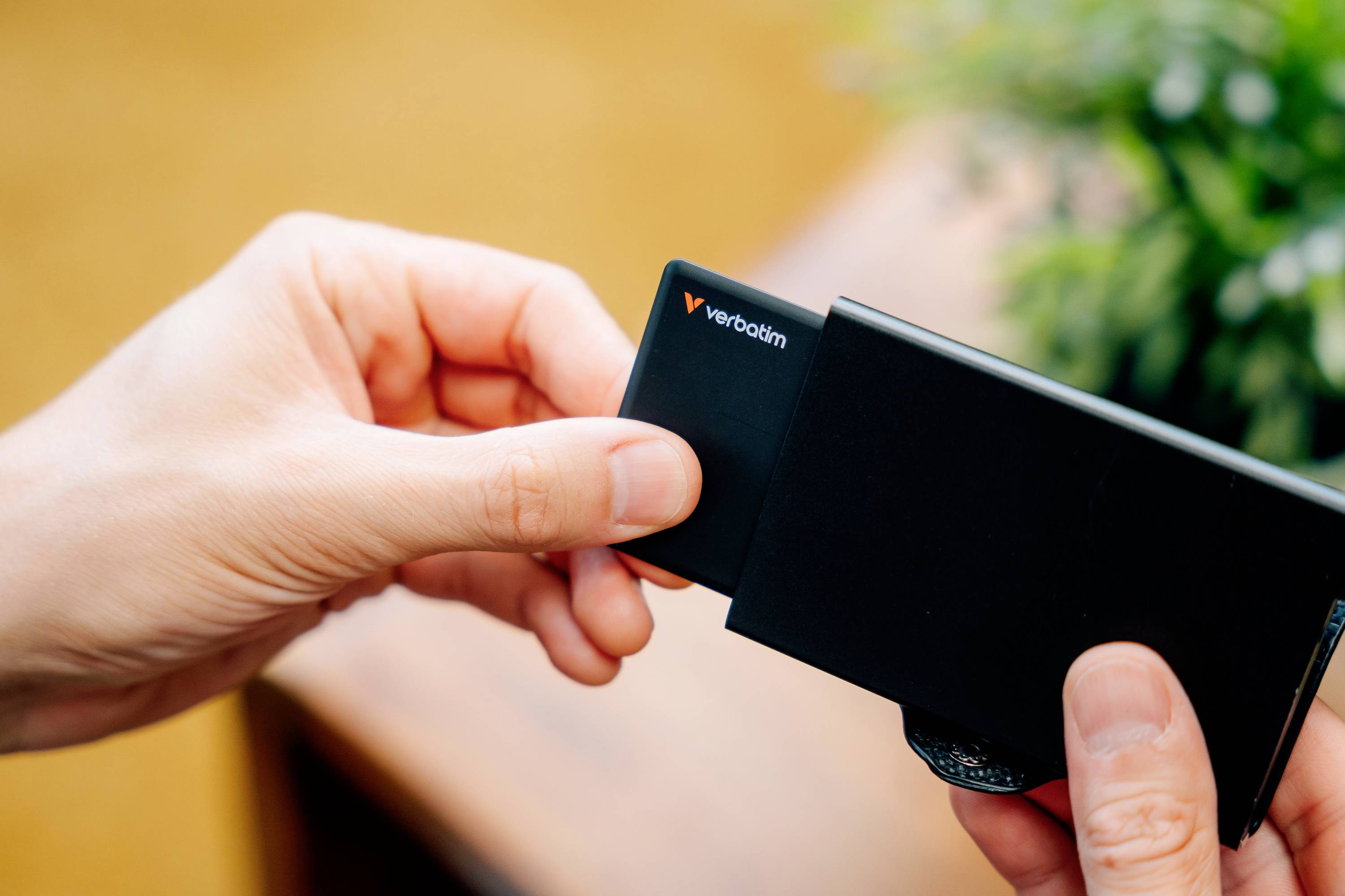 A person is holding a black Verbatim memory card and inserting it into a black device in an indoor setting.