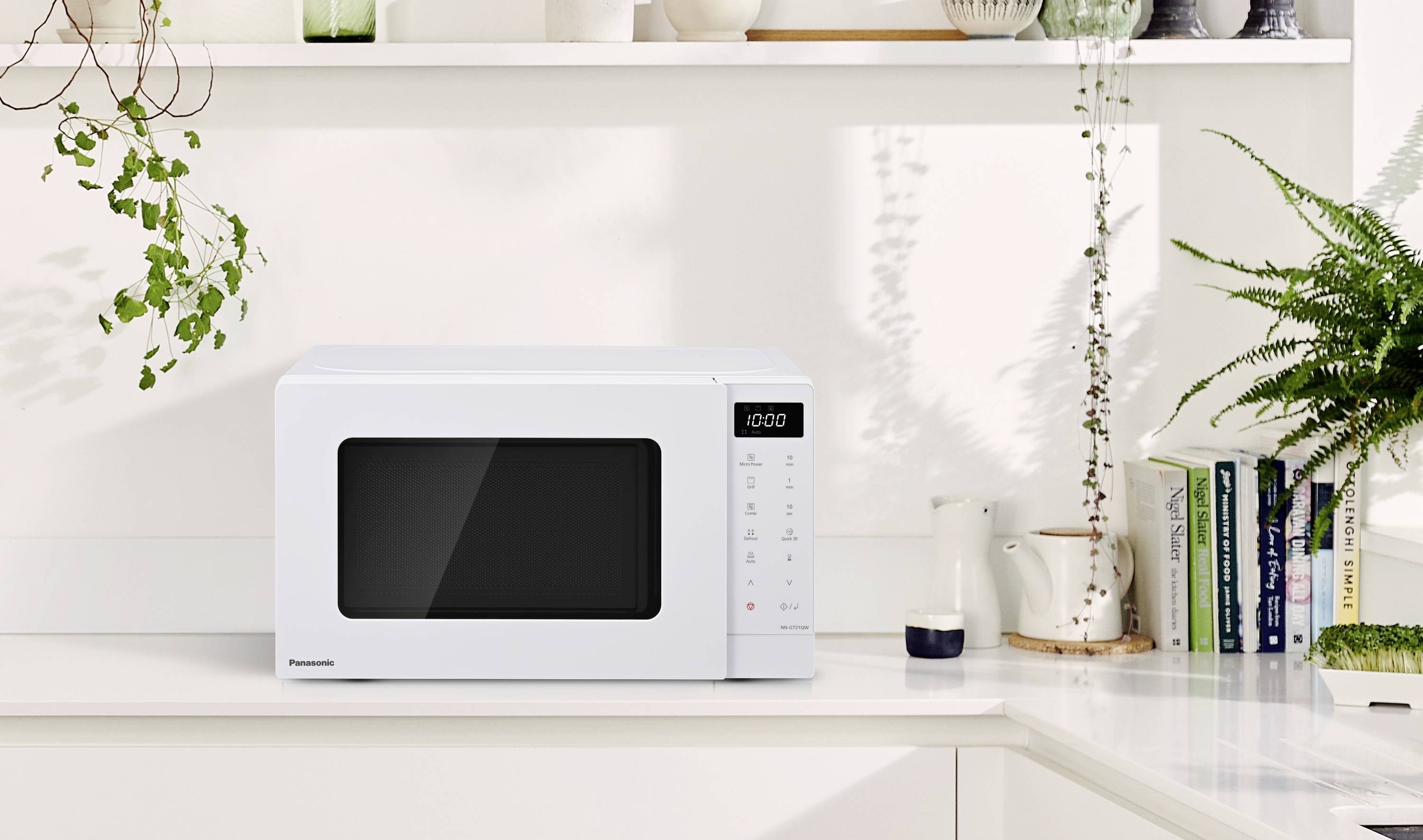 White microwave on a kitchen worktop, surrounded by plants and books, shows 4:00 on the digital display.
