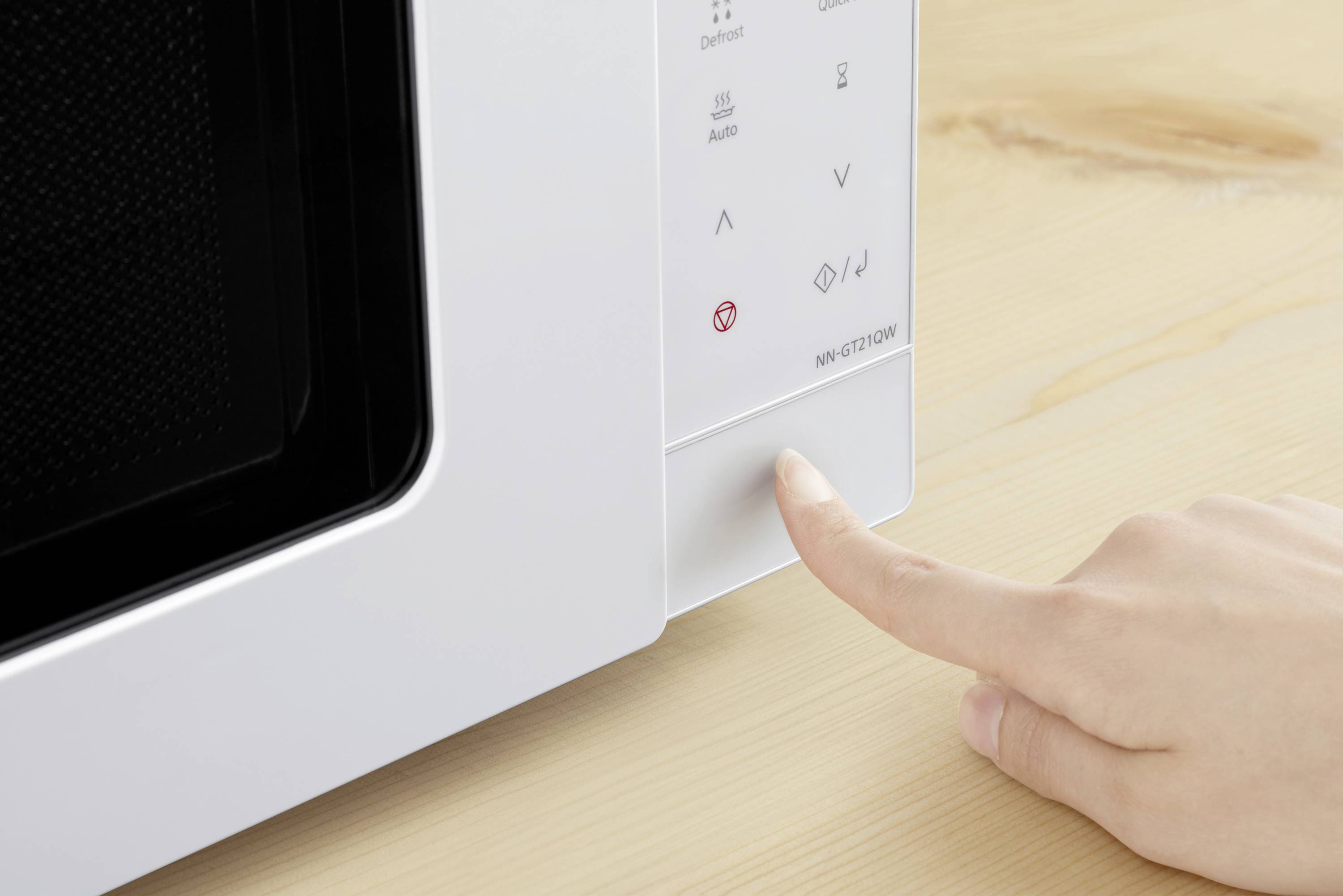 A hand presses the start button of a white microwave on a wooden table.