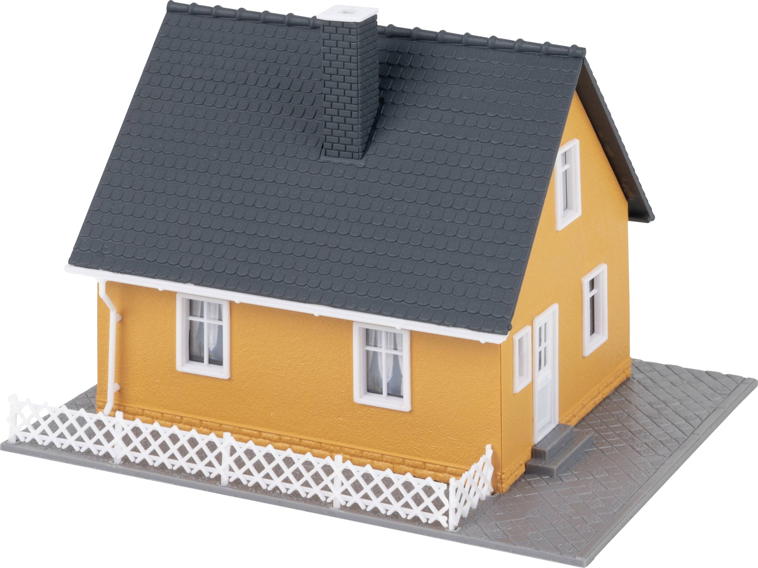A small model house with an orange facade, black roof tiles, and white windows, surrounded by a white picket fence.