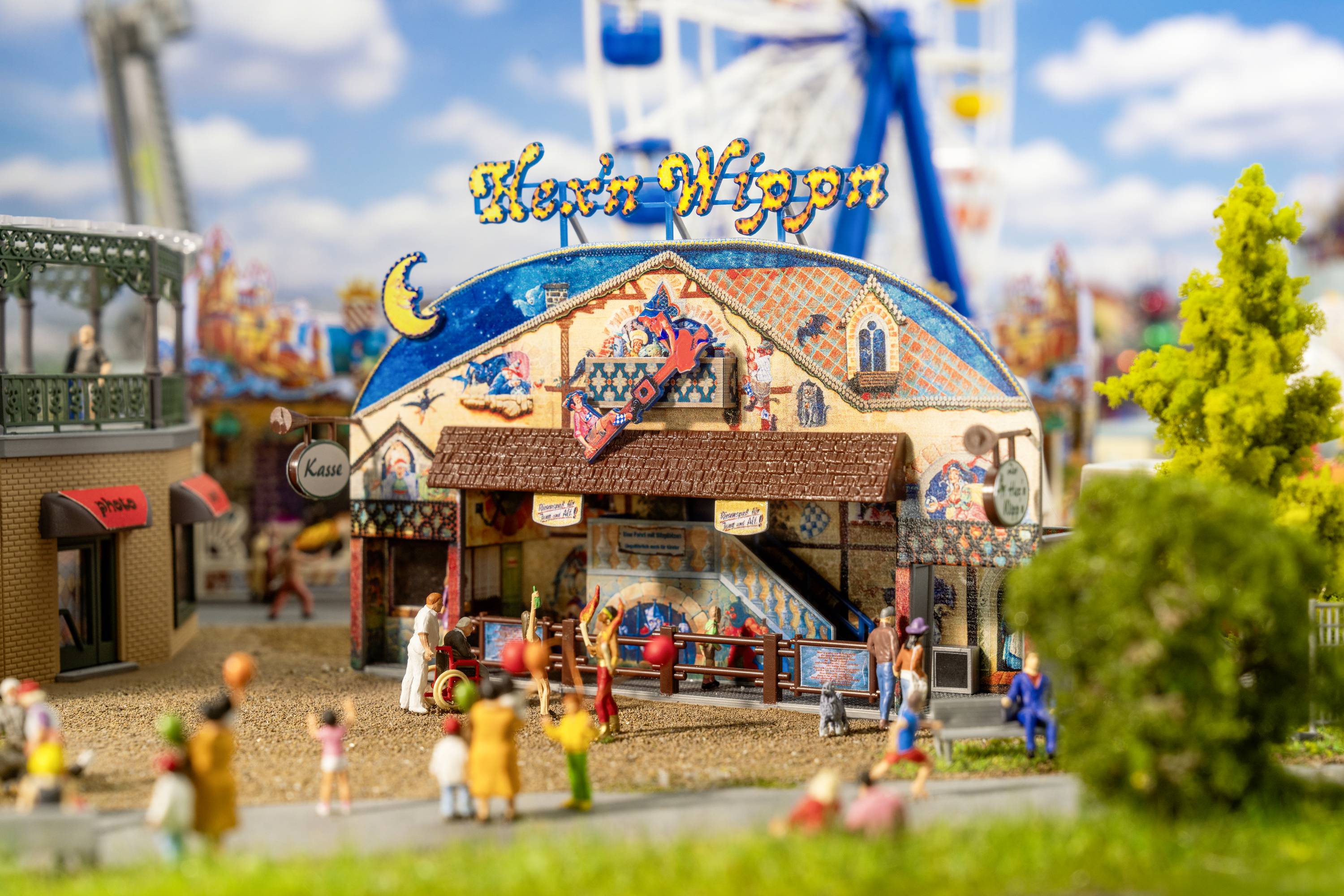 A miniature fairground scene with a colourful 'Witches' Wagon' tent, surrounded by figures and trees, against a blue sky with clouds.